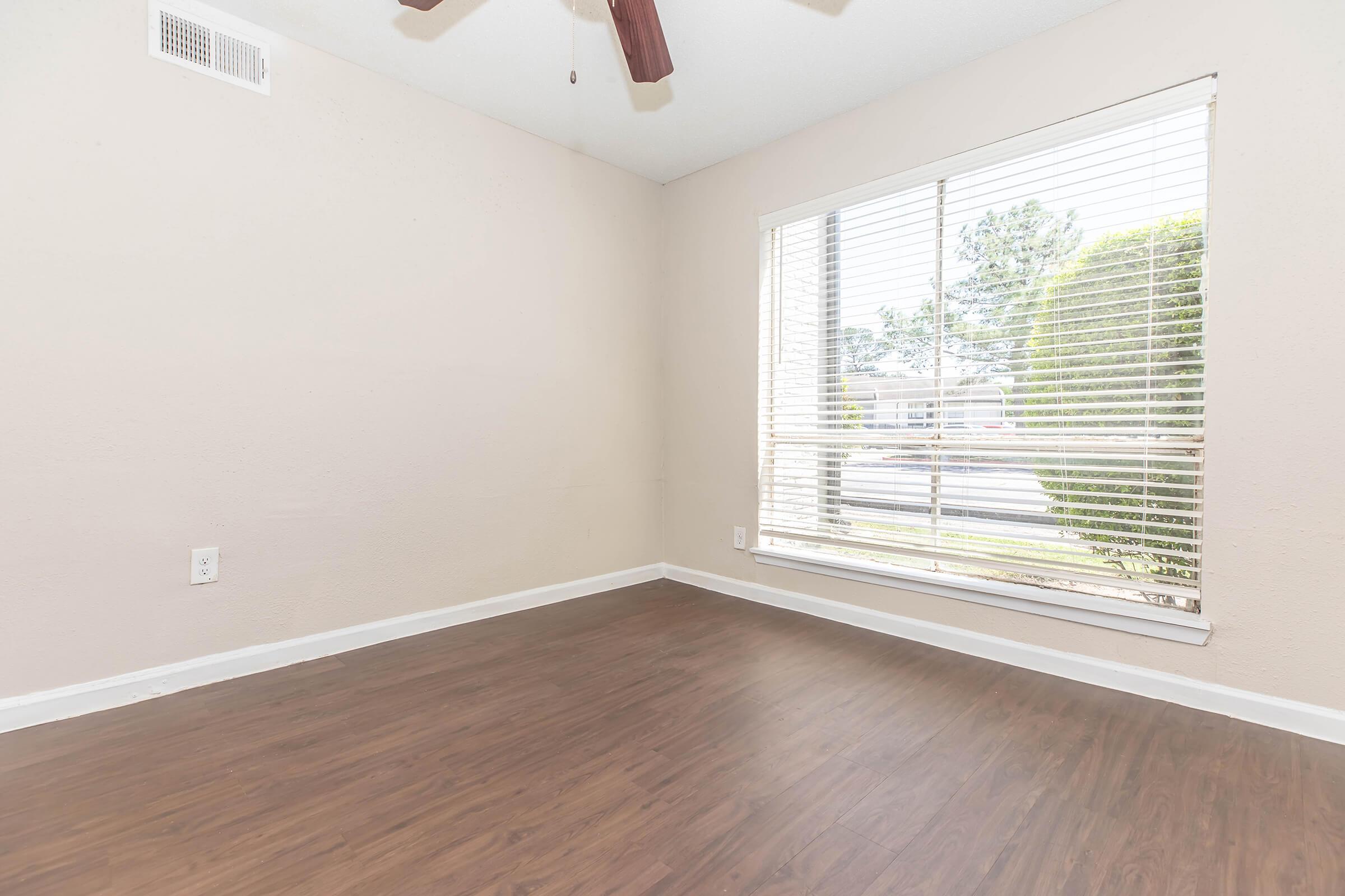 Empty room with light-colored walls and a wooden floor, featuring a ceiling fan and a large window with blinds. The window allows natural light to enter, illuminating the space. The exterior is visible through the window, with greenery outside.