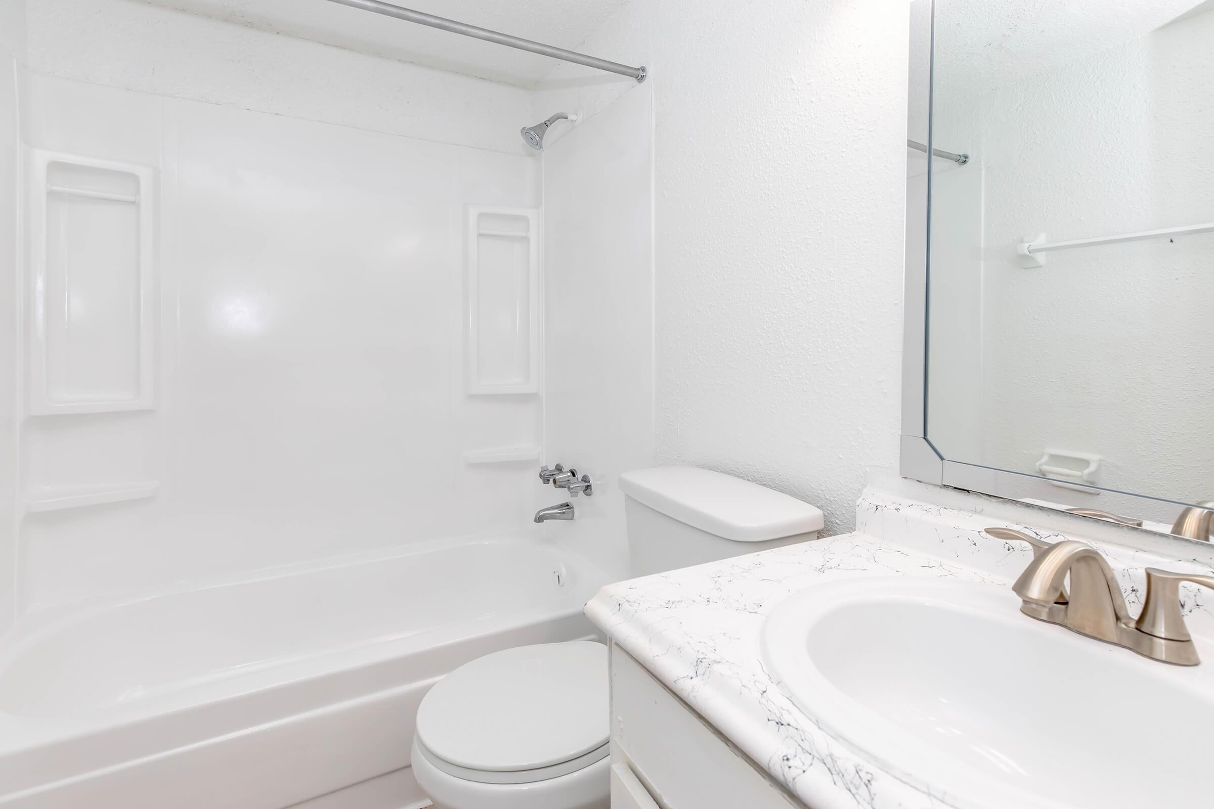 A clean, modern bathroom featuring a white bathtub and shower combination, a toilet, and a sink with a marble countertop. The walls are light-colored, and there is a mirror above the sink. The overall design is simple and functional, reflecting a tidy and bright space.