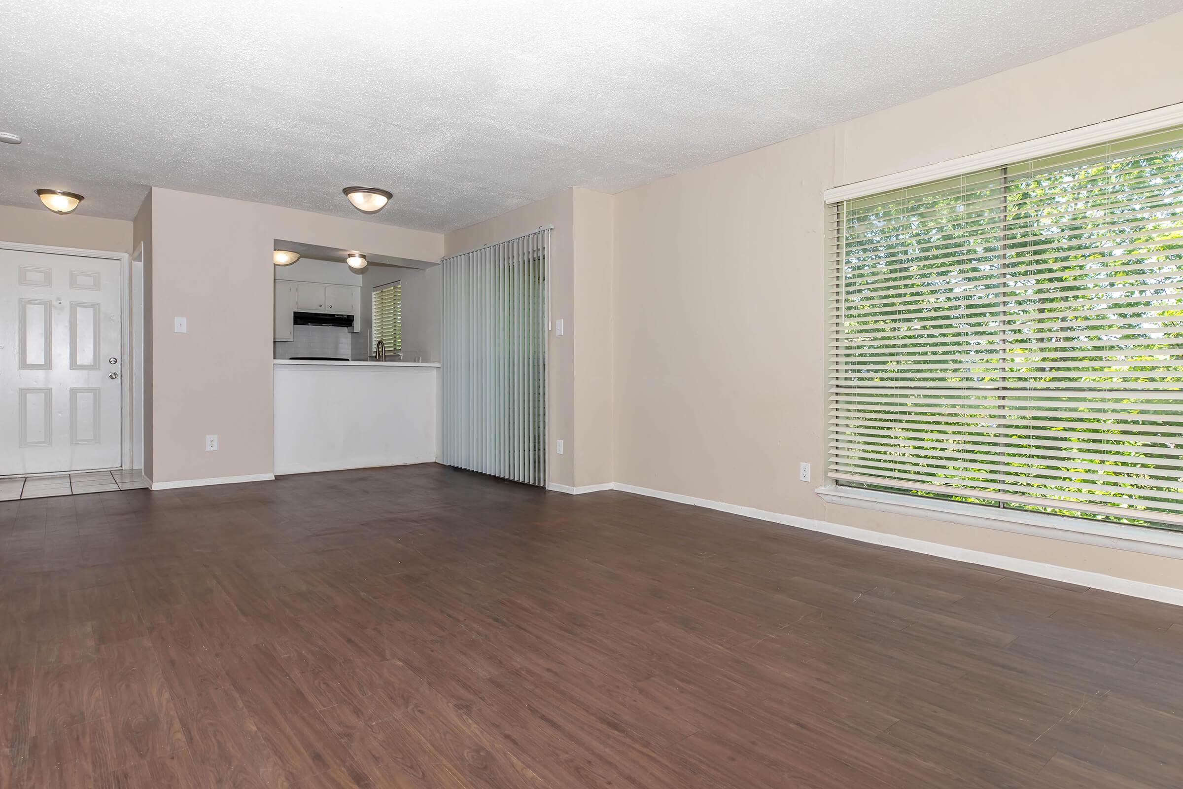A spacious living area with hardwood-like flooring, featuring large windows with blinds allowing natural light. The room leads to a kitchen area, partially visible through an open entry. The walls are painted in a neutral color, and there is a front door visible on the left.