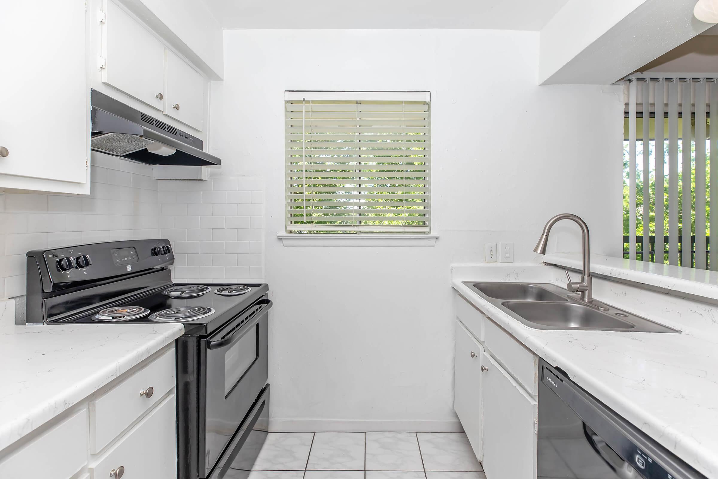 A modern kitchen featuring white cabinetry, a black stove and oven, a stainless steel sink, and a countertop. There is a window with blinds that allows natural light. The flooring is tiled in a light color, and the overall design is clean and minimalist.