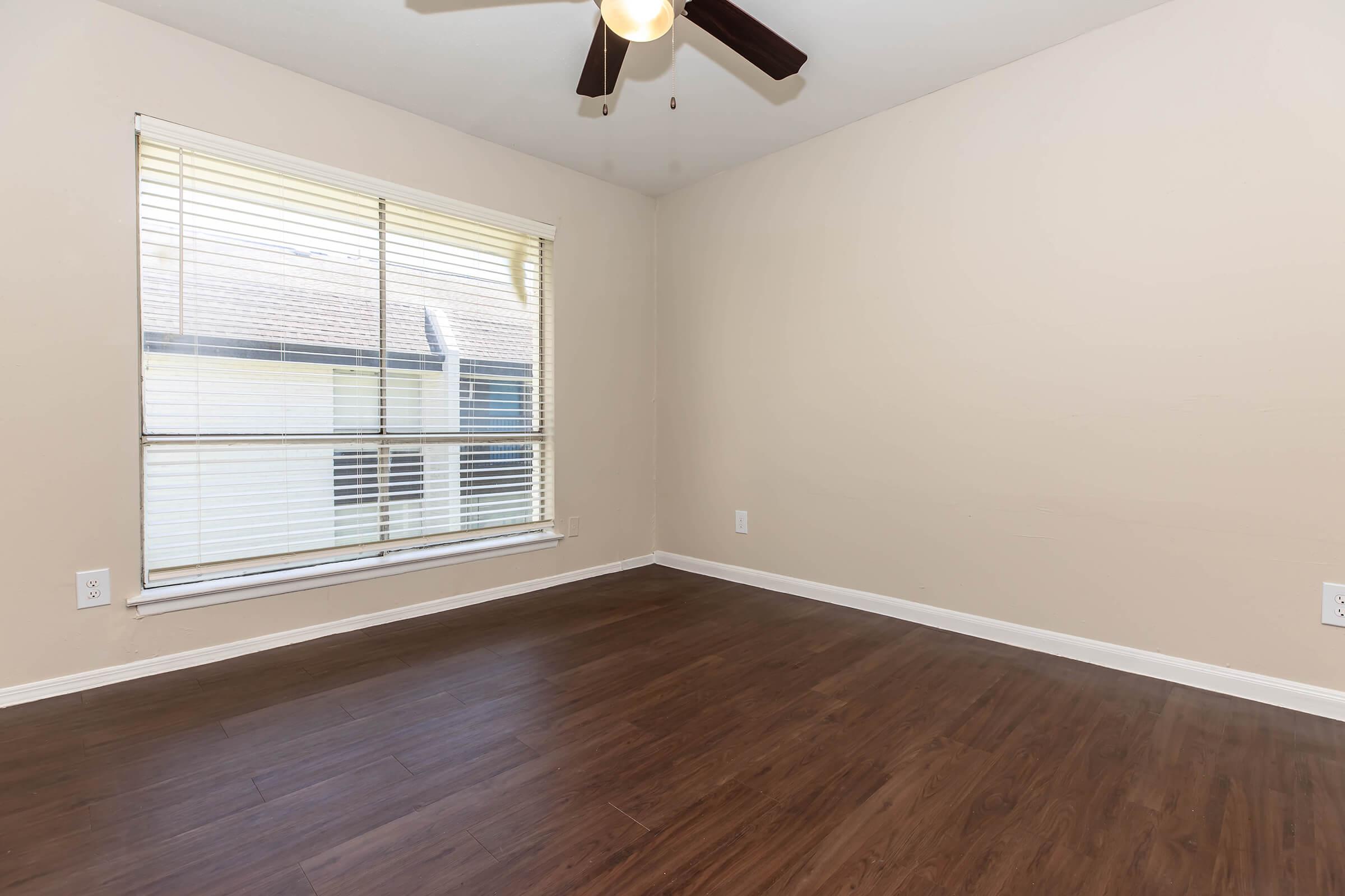 Empty room with light brown walls and a ceiling fan. There is a large window with blinds letting in natural light, and the floor is wooden. The space is uncluttered, providing a blank canvas for potential furnishings or decoration.