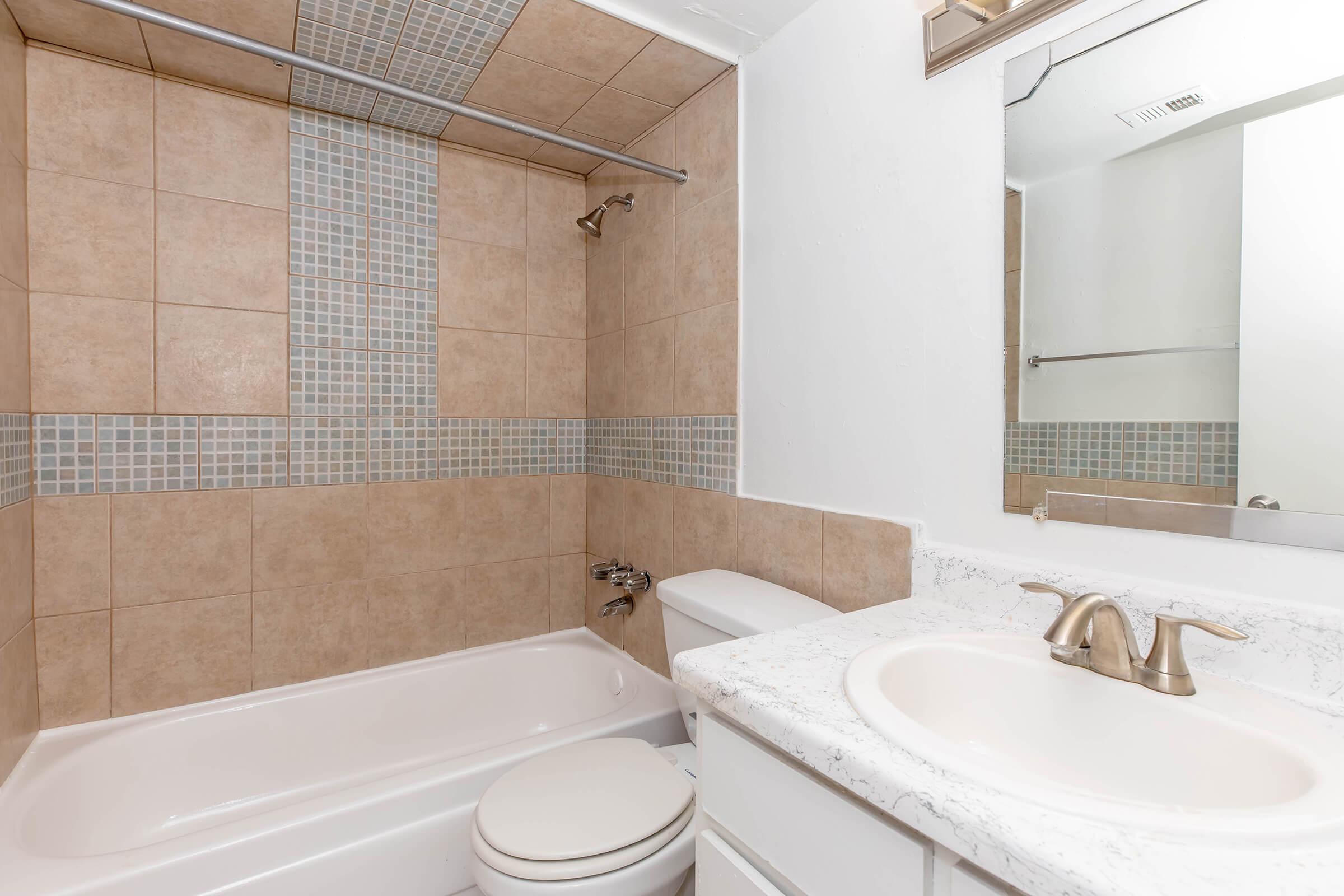 A clean, modern bathroom featuring a bathtub with beige tiled walls, a showerhead, a white toilet, and a sink with a marble countertop. A large mirror reflects the space, and small square tiles add a decorative touch to the wall above the bathtub.