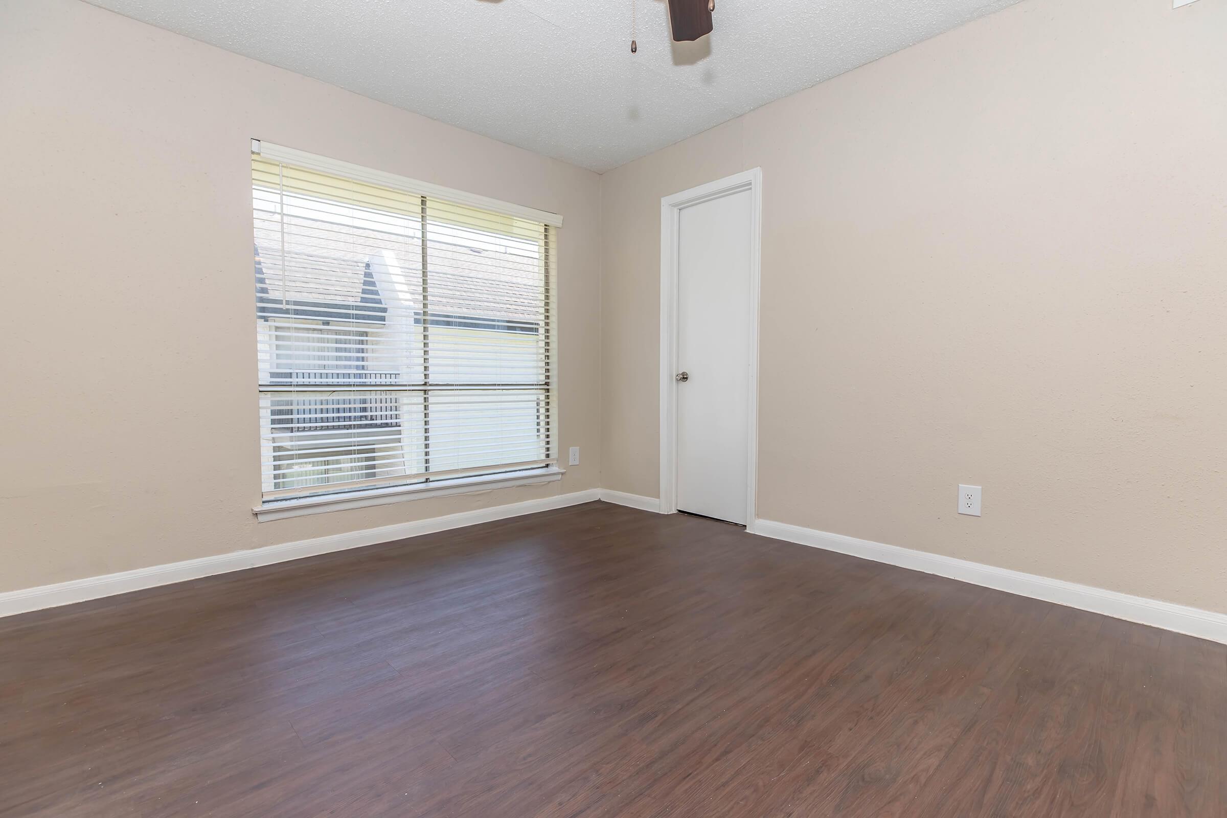 Empty room with light-colored walls and a wooden floor. A window with blinds allows natural light to enter, and a closed white door is visible on one side. The space is well-lit and appears simple and minimalistic.
