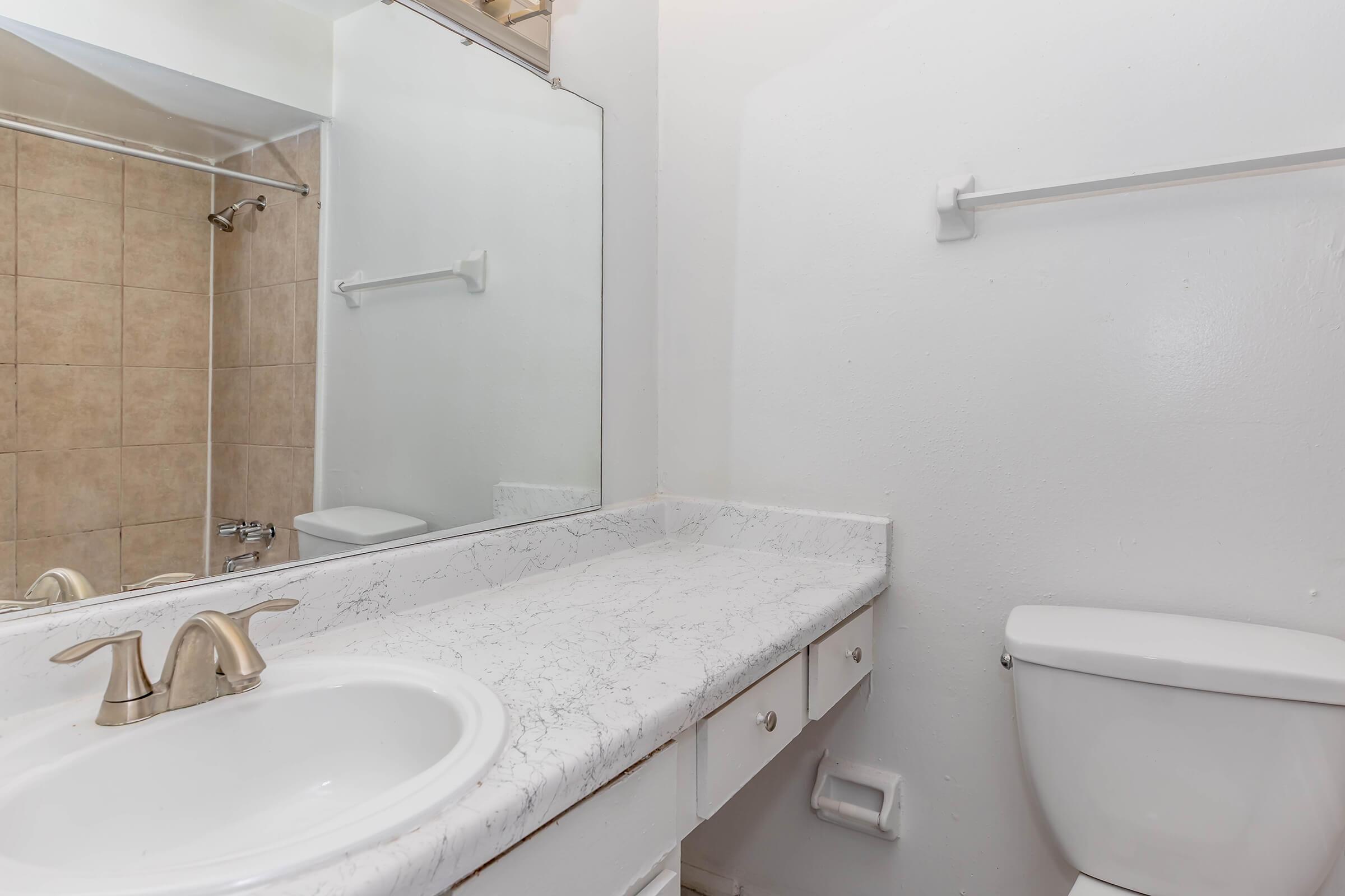 A clean bathroom with a white countertop, a round sink with gold faucets, a large mirror above the sink, a tiled shower area visible in the background, and a white toilet. The walls are painted white.