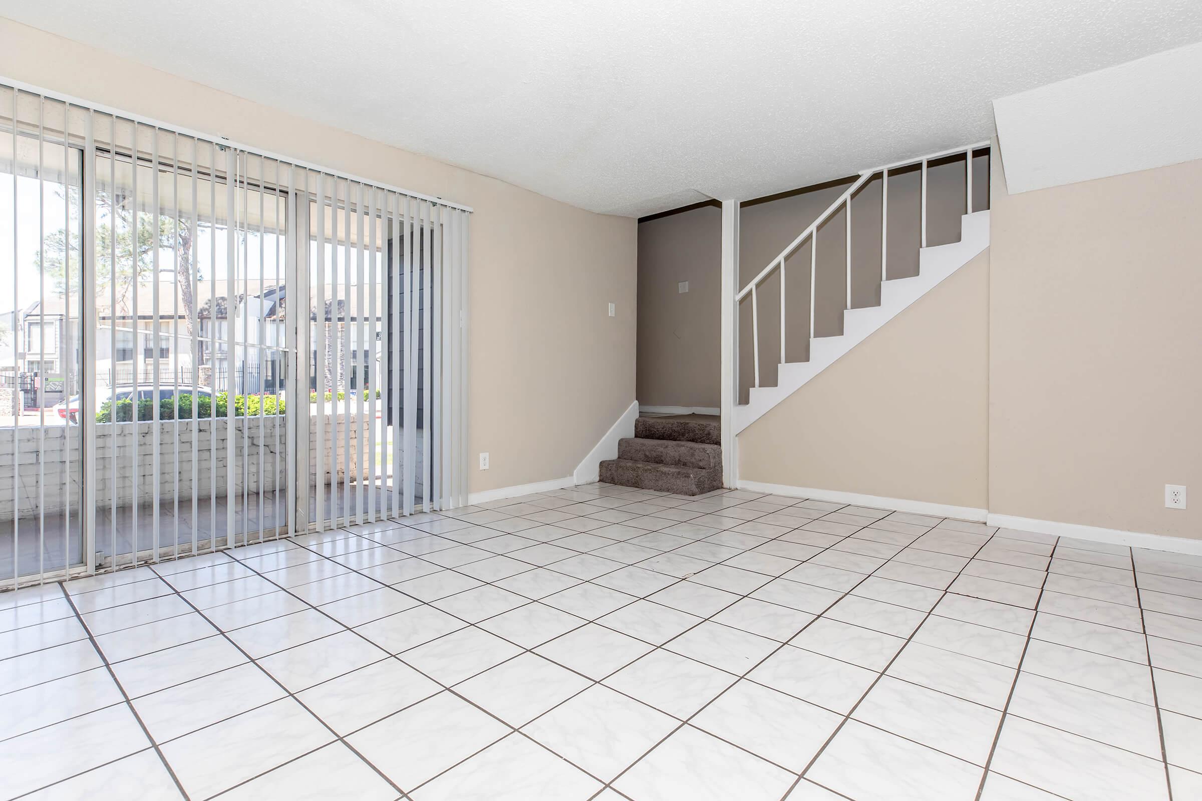 Spacious, empty living room with tiled flooring and large sliding glass doors leading to an outdoor area. A staircase is visible in the background, along with light-colored walls. The room is bright and airy, providing an inviting atmosphere.