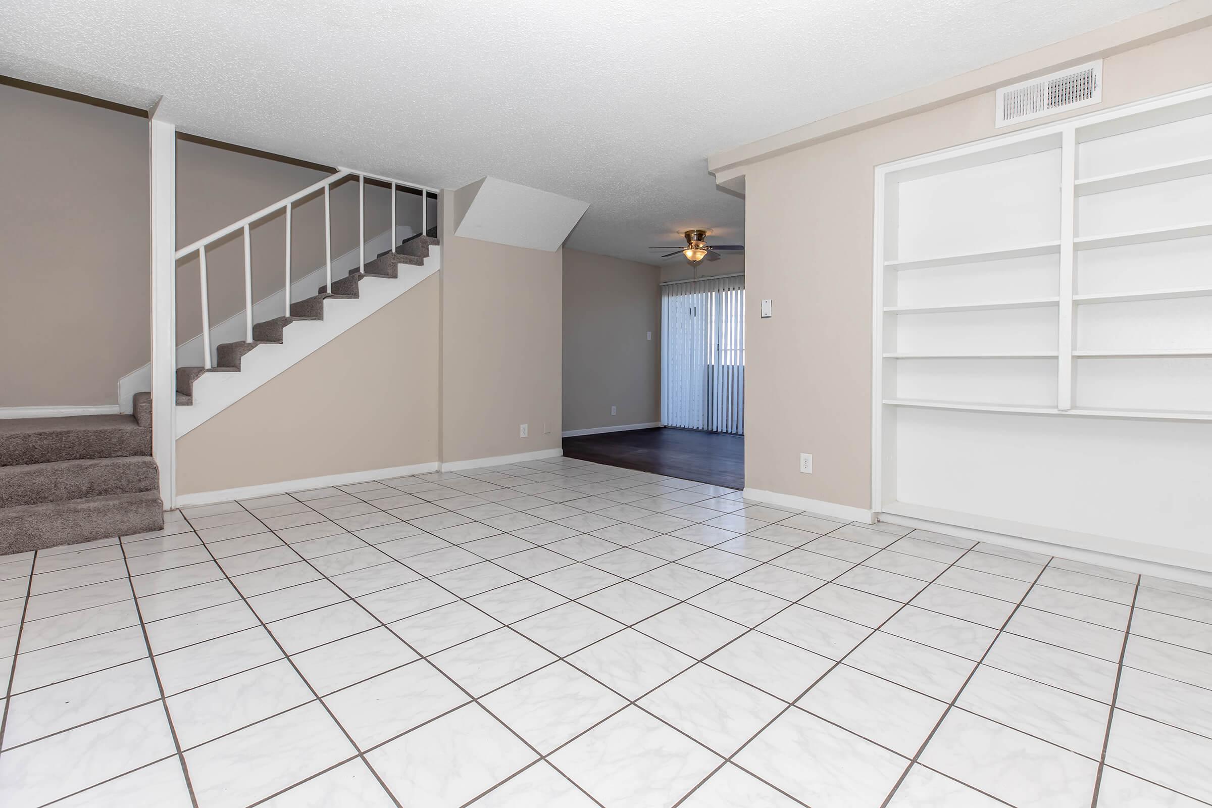 A spacious, empty living area with white tile flooring, light beige walls, and a staircase on the left. The room features a ceiling fan and a built-in shelf on the right. A sliding glass door leads to an outdoor space, enhancing natural light in the room.