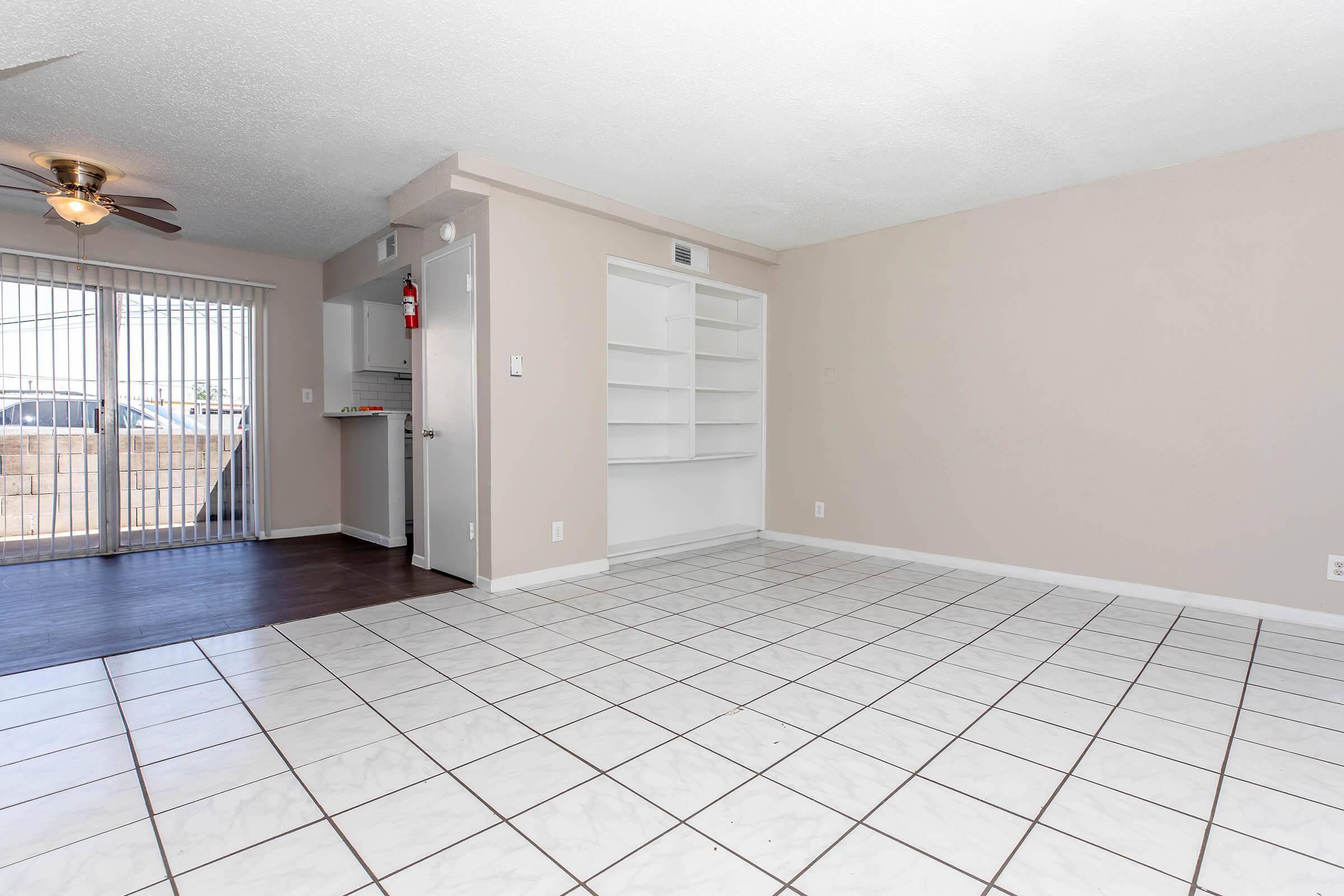 A spacious, empty room featuring tiled flooring, a ceiling fan, and a large window with vertical blinds letting in natural light. There's a door leading to another area and a built-in shelf along one wall. The walls are painted in a light color, giving the room a bright and airy feel.