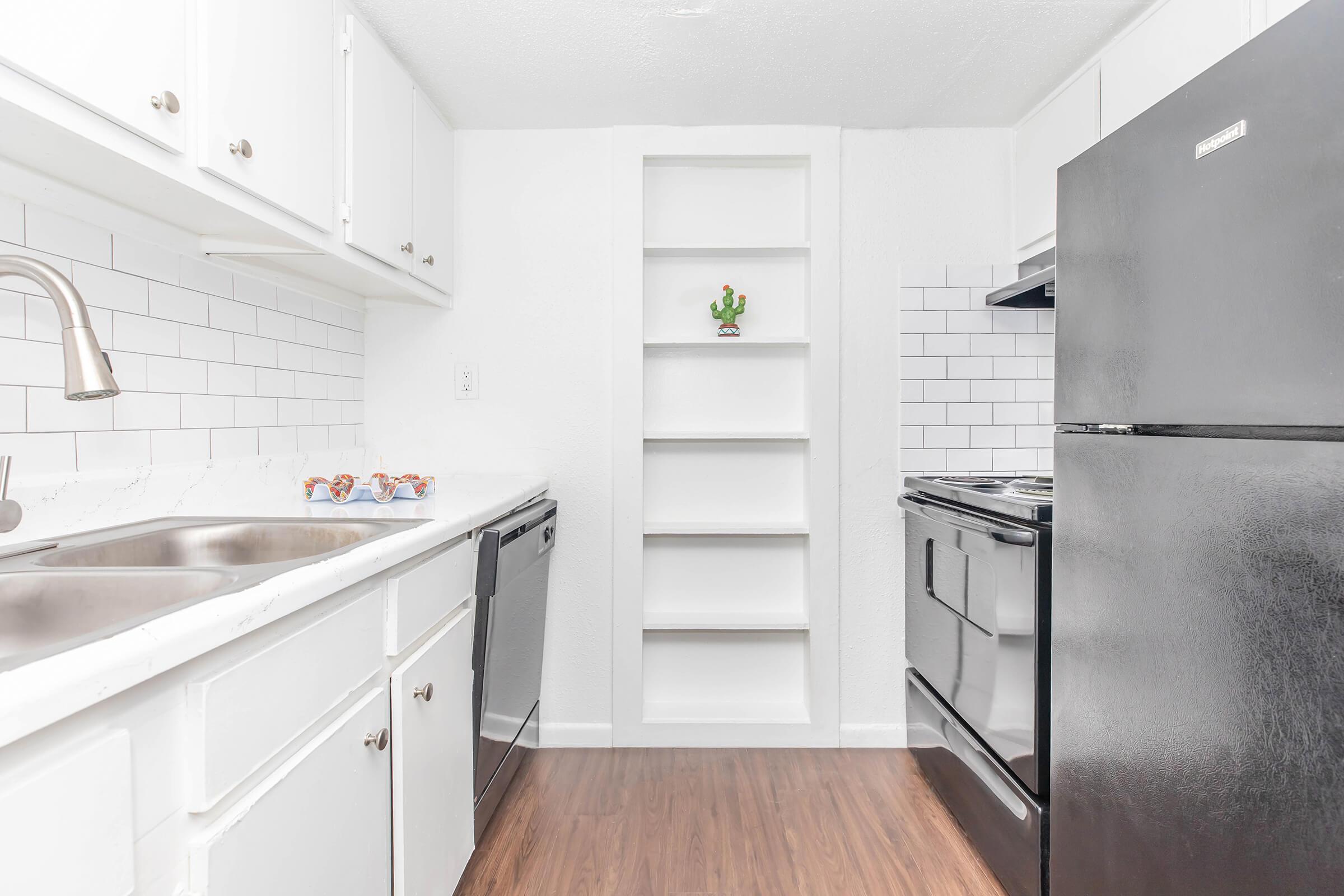 A modern kitchen featuring white cabinets, a stainless steel sink, a black refrigerator, and an oven. There's a dishwasher with a black front, and open shelves are built into one wall. The floor is covered in brown laminate, and small decor items are placed near the sink. Light-colored walls provide a bright atmosphere.