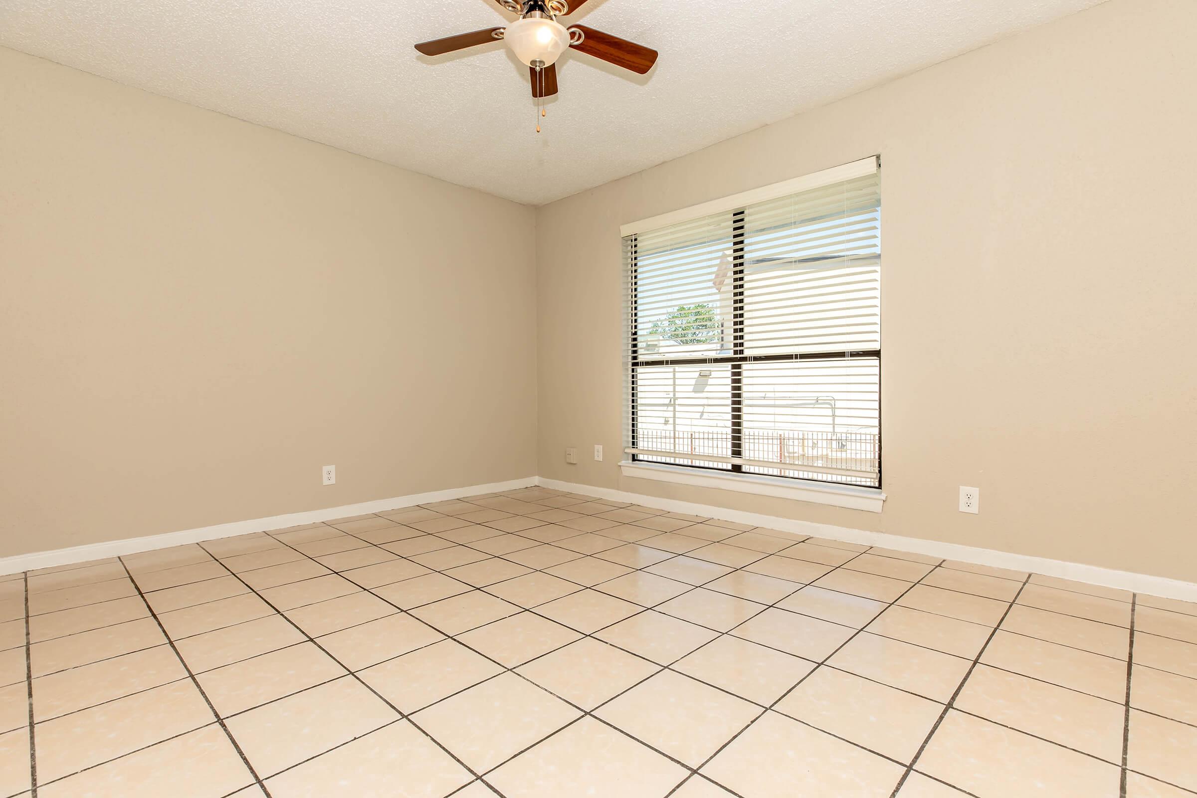 A spacious, empty room with light beige walls and glossy cream-colored tile flooring. There is a window with blinds allowing natural light, and a ceiling fan with wooden blades is installed. The room has a clean, uncluttered appearance, suitable for various uses.