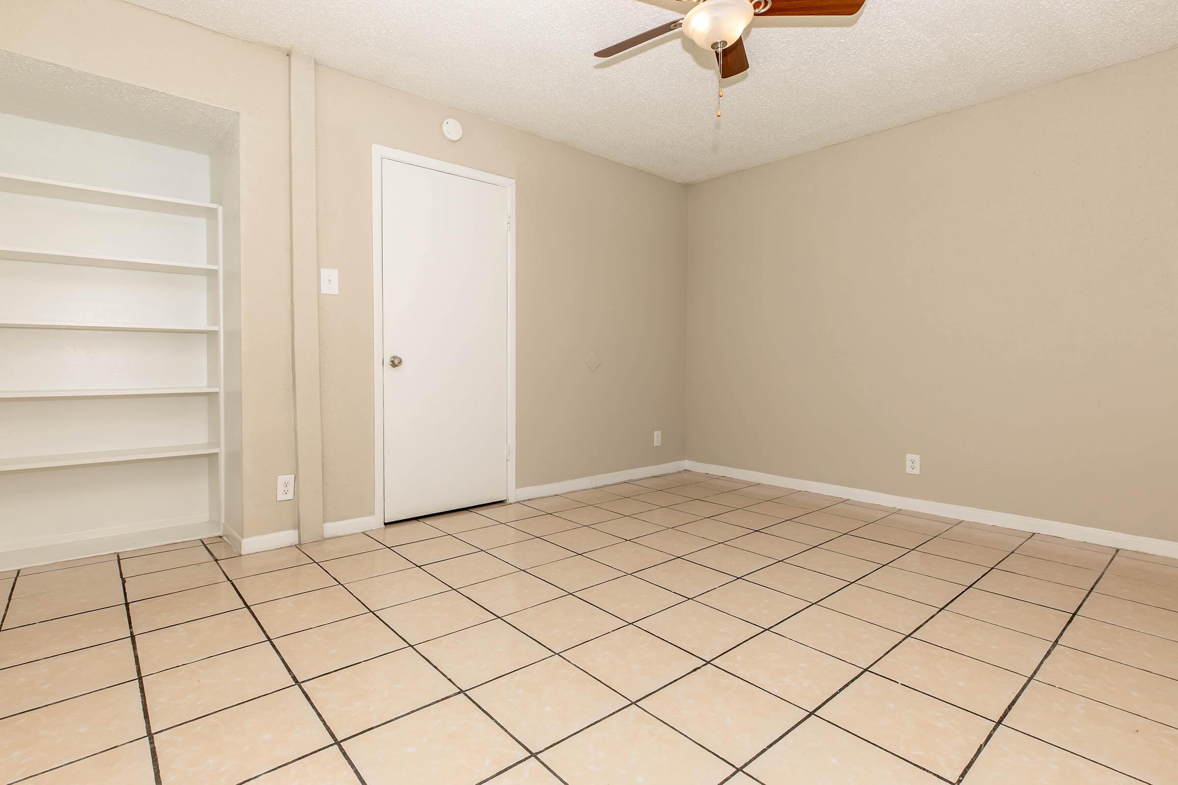 A clean, empty room with beige walls and tiled flooring. There's a ceiling fan, a closed white door, and a built-in bookshelf on one side. The space is well-lit and features a simple, uncluttered design, suitable for various uses or decorations.