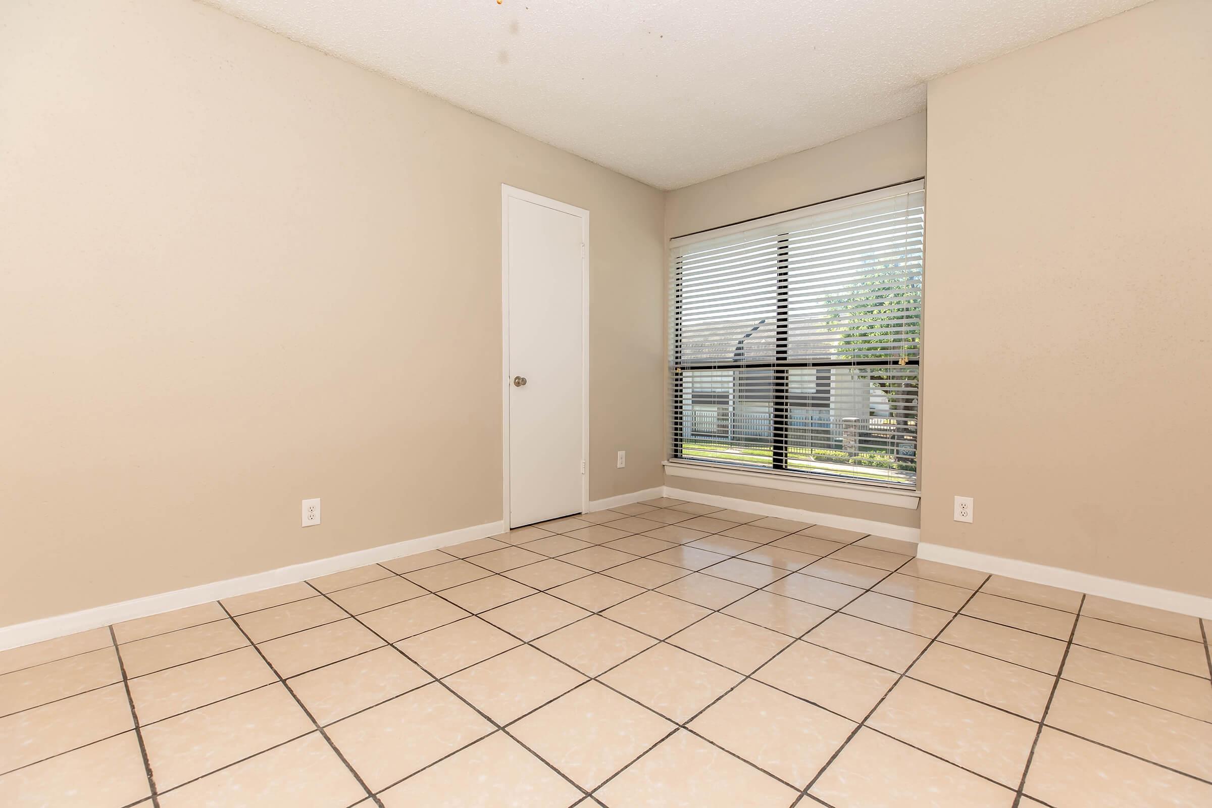 Empty room with beige walls and a tiled floor. A closed white door is on the left, and a large window with horizontal blinds overlooks a green outdoor area. The room appears well-lit and spacious, suitable for various uses.
