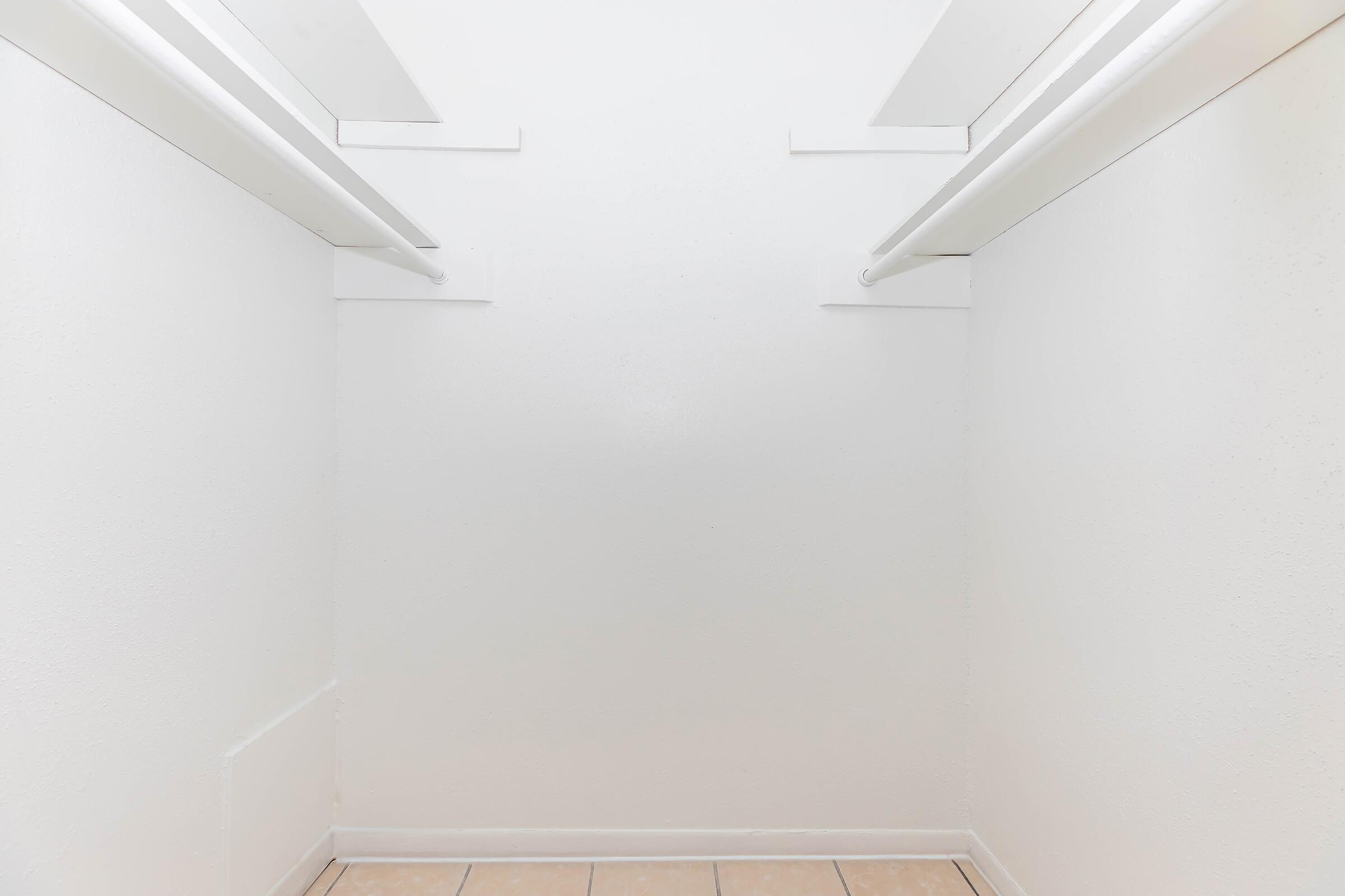 Empty closet with two horizontal hanging rods on opposite sides, and plain white walls. The floor is tiled with light-colored tiles, creating a minimalistic and uncluttered appearance.
