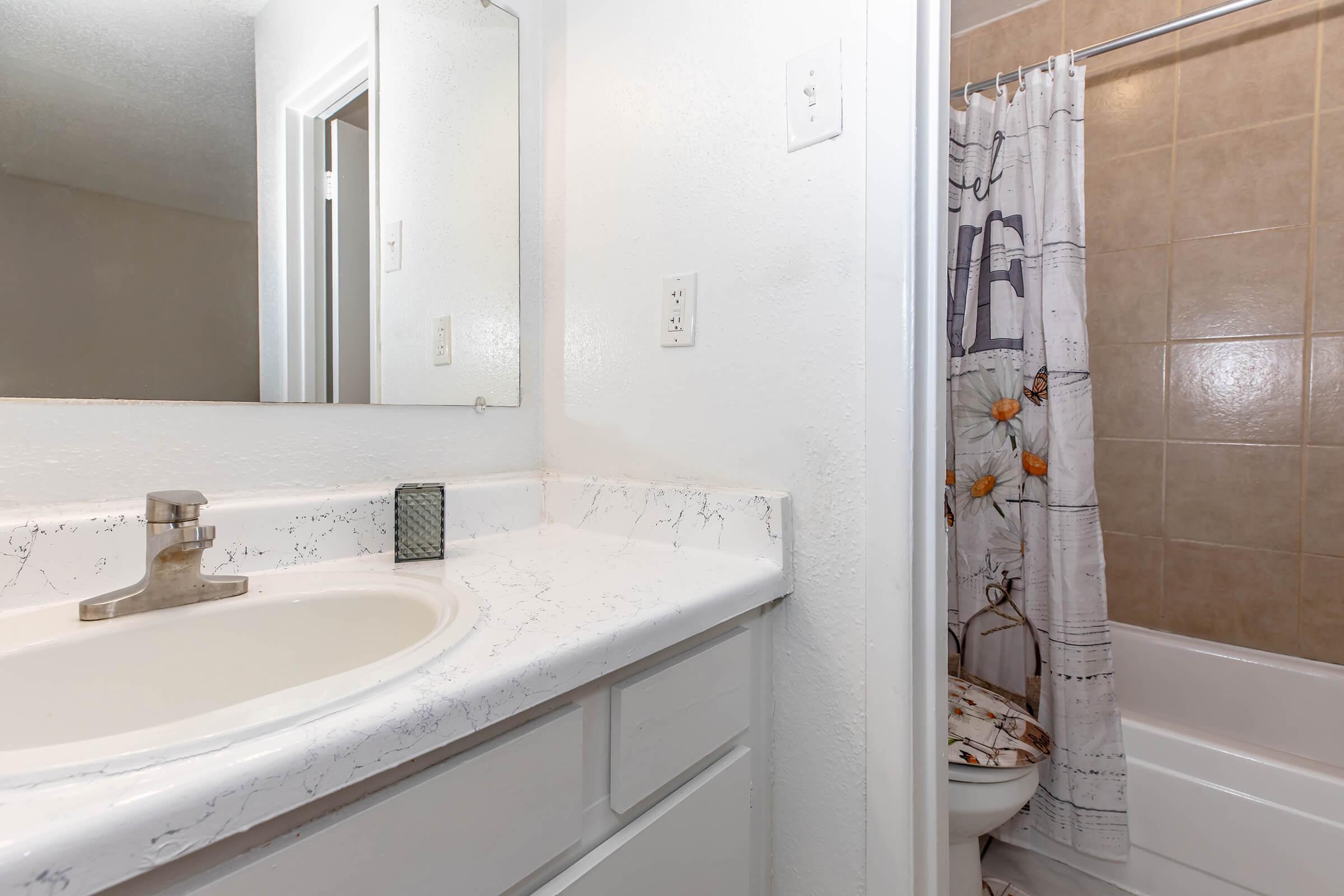 A clean bathroom featuring a white countertop with a sink, a large mirror on the wall, and a bathtub with a decorative shower curtain. The walls are painted white, and the flooring has neutral-colored tiles. There is a towel rack next to the shower.