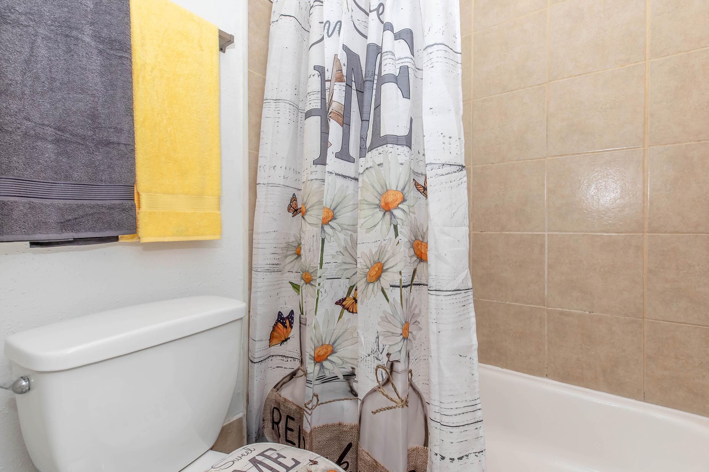 A bathroom featuring a shower with a floral curtain adorned with daisies and butterflies. There is a white toilet beside the shower, and two towels—one yellow and one gray—hang on the wall. The tile walls are light beige, creating a warm atmosphere.