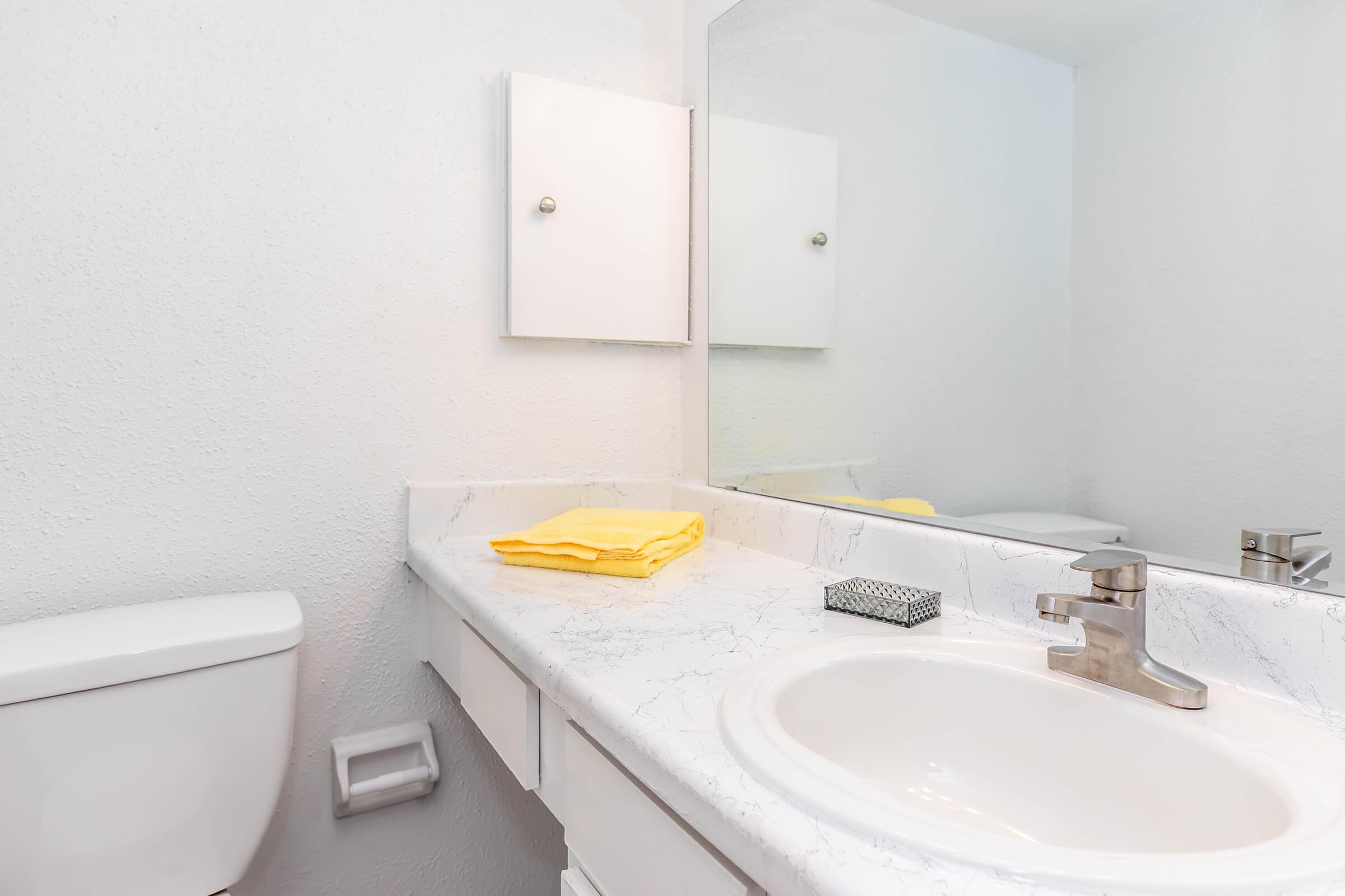 A clean bathroom featuring a white countertop with a sink, a faucet, a small black storage box, and neatly folded yellow towels. A toilet is visible on the left, and there is a simple mirrored cabinet above the sink. The walls and surfaces are in neutral tones, creating a bright and airy atmosphere.