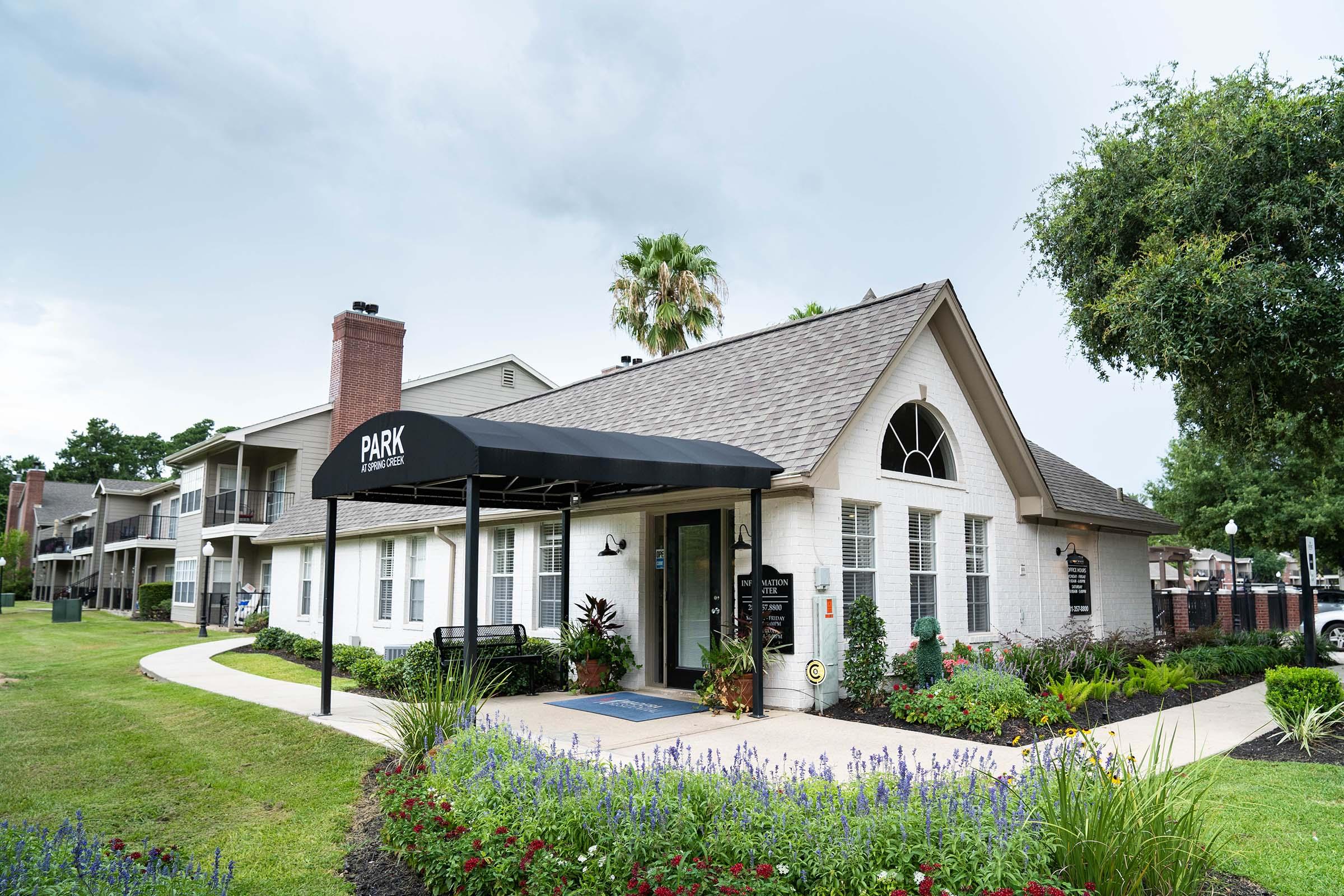 A well-maintained building with a sloped roof and an awning that reads "PARK." Accessible pathways lead to the entrance, surrounded by greenery and flower beds. The landscape features trees and shrubs, providing a pleasant outdoor environment. Overcast sky in the background hints at possible rain.