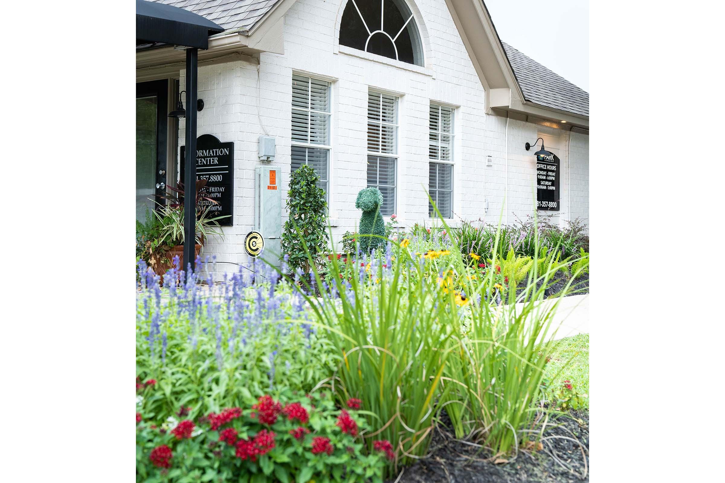 A well-kept building entrance with white brick walls, featuring large windows and a decorative arch. Lush green plants and colorful flowers, including red and purple blooms, surround the entrance, creating a welcoming atmosphere. Signs indicate it is an information center.
