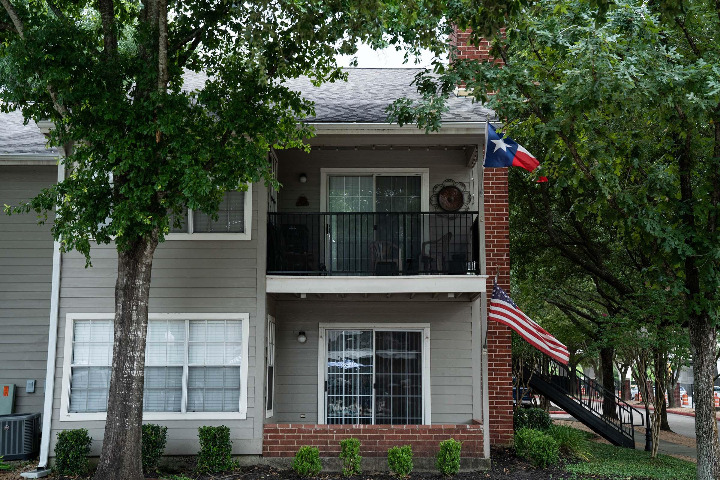 A two-story apartment building with a balcony featuring a Texas flag and an American flag. The surrounding area has trees and neatly trimmed bushes, providing a natural setting. Windows are adorned with curtains, and a staircase is visible in the background, leading to the upper floor.