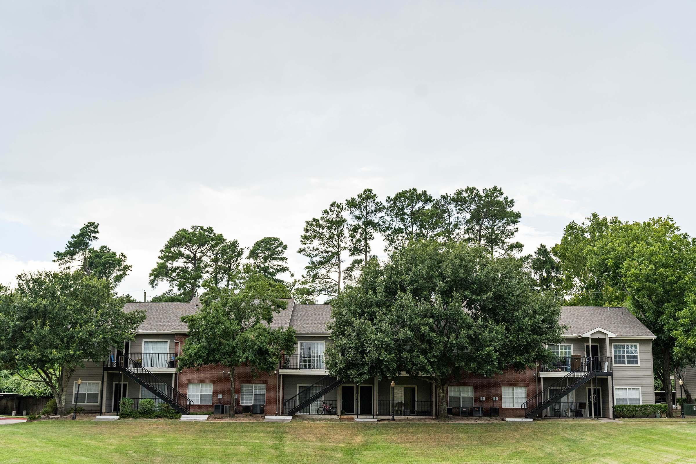 A two-story apartment building with brick exterior and balconies, surrounded by lush green trees and grass. The sky is overcast, suggesting possible rain. The scene reflects a calm residential environment.
