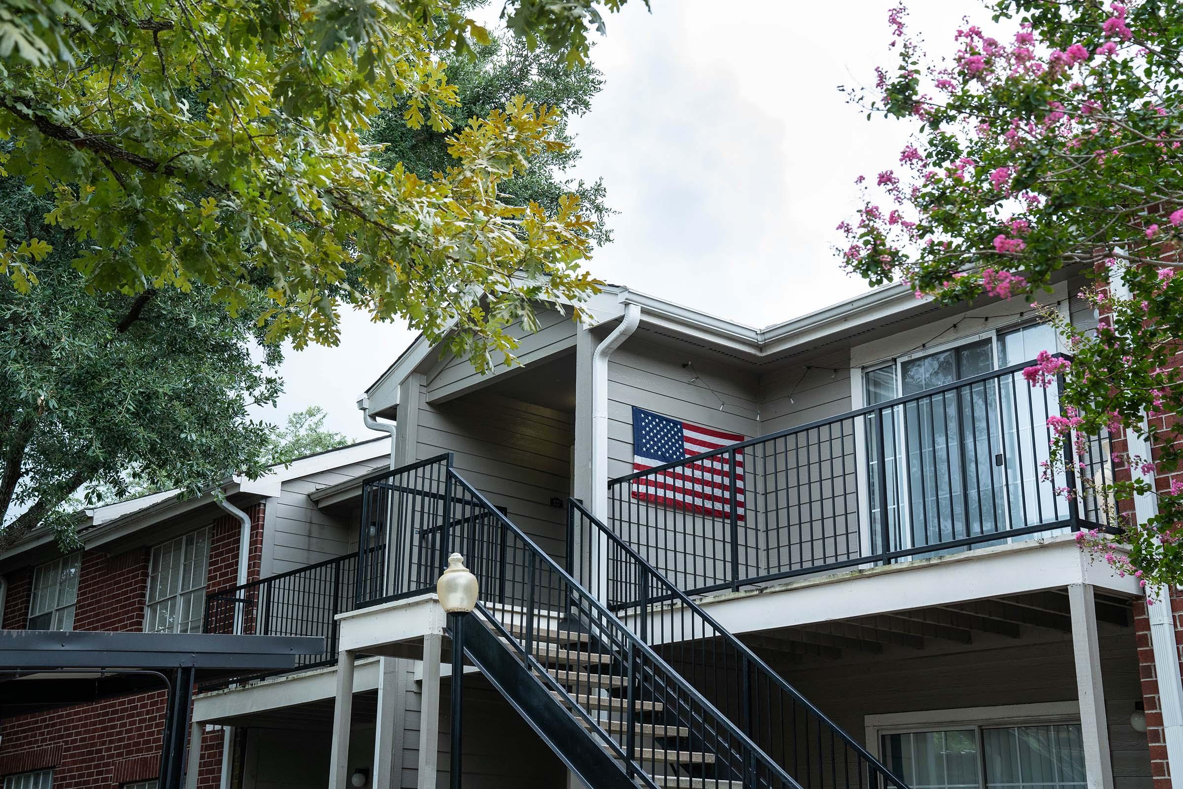 A multi-story apartment building featuring a staircase leading to an upper balcony. An American flag hangs on the balcony wall, surrounded by green trees and pink flowering plants. The building has a modern architectural style with large windows and a neutral color palette.