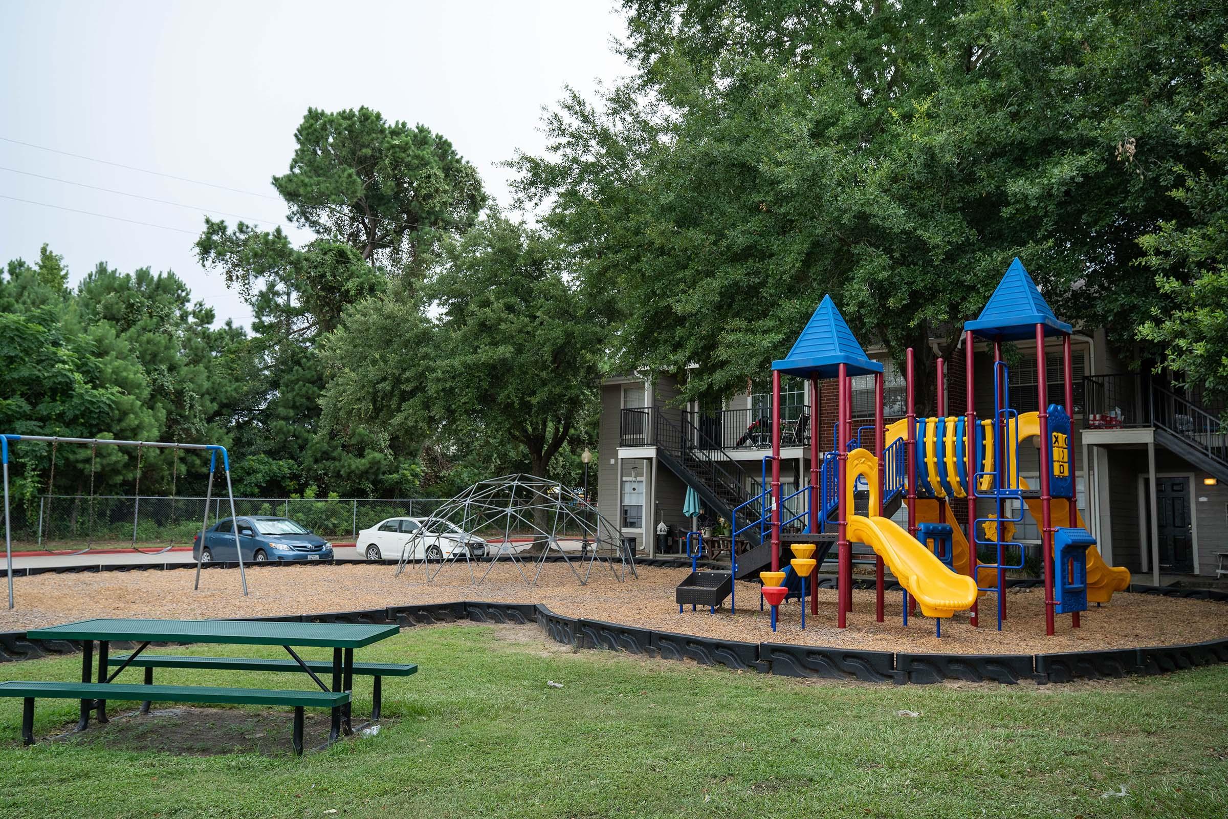 A playground featuring colorful slides and climbing structures surrounded by grassy areas. Nearby, there is a swing set and a geodesic climbing dome. In the background, there's a parking area with a car and a multi-story building visible. Tall trees provide shade around the playground.