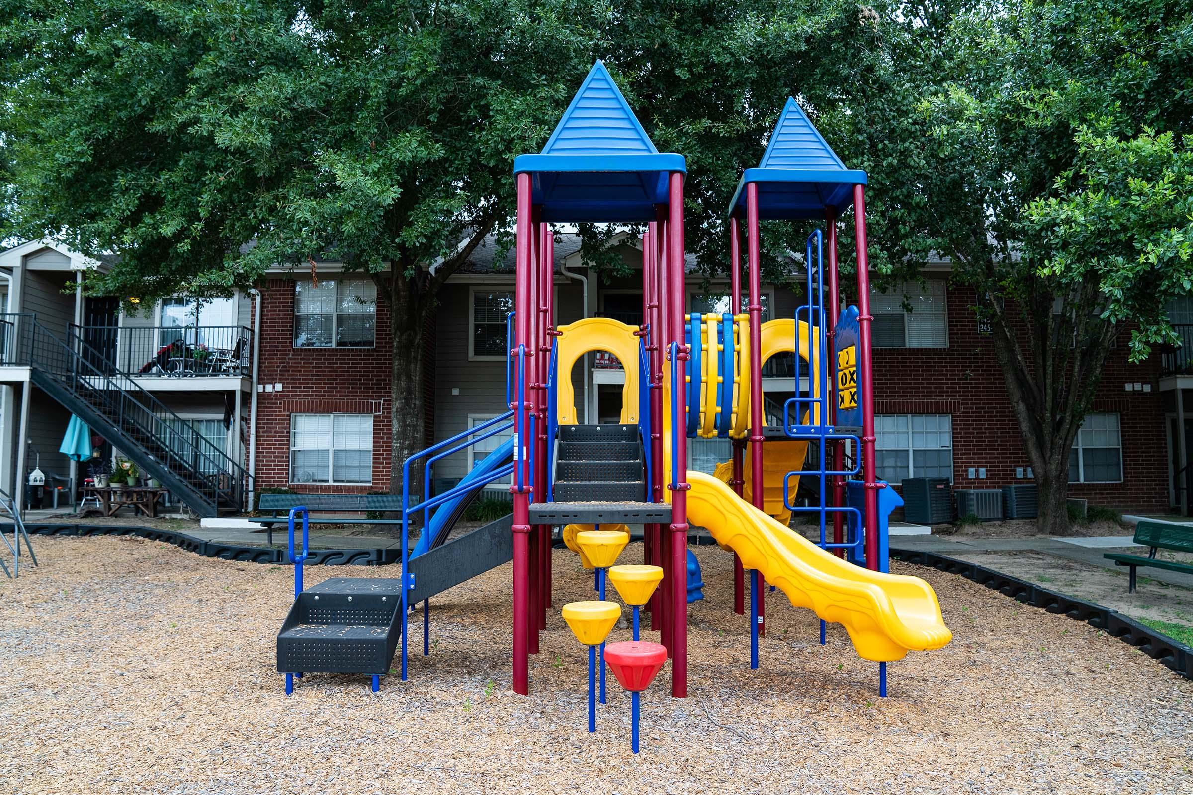 Colorful playground equipment featuring two slides, climbing structures, and steps, surrounded by a gravel play area. It is situated near a residential apartment complex with green trees in the background.