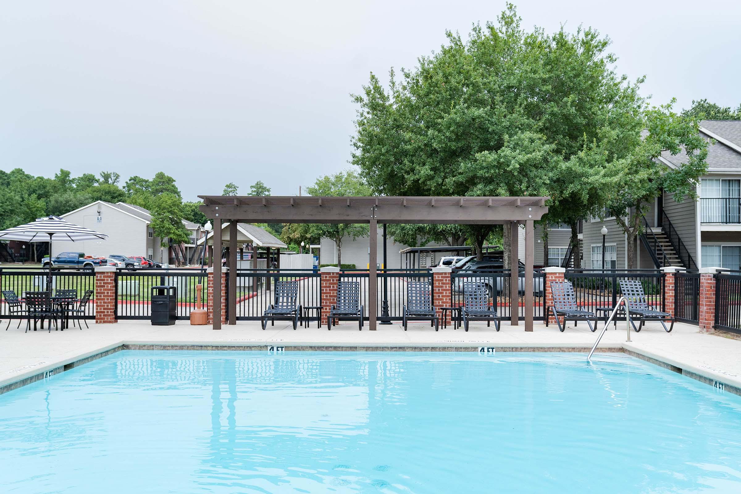 A clear swimming pool with a wooden pergola beside it, surrounded by lounge chairs. In the background, there are apartment buildings and a large tree, with parked cars visible. The scene is calm, likely set in a residential area. The sky is overcast, hinting at possible rain.