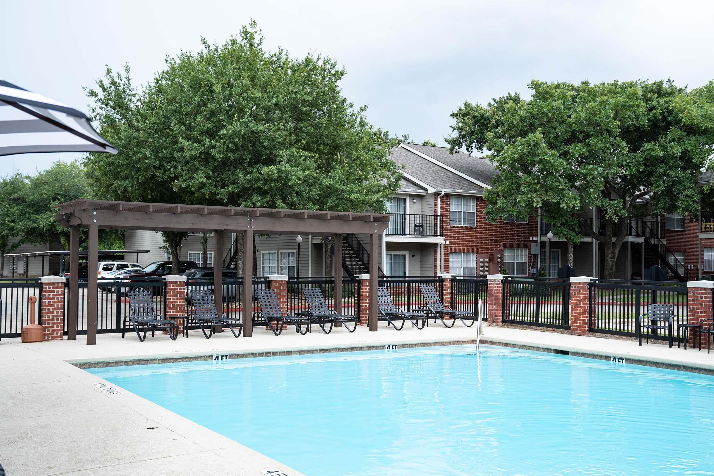 A clear blue swimming pool surrounded by a fenced area. There are several lounge chairs arranged nearby, and a pergola structure with a roof in the foreground. In the background, there are red-brick apartment buildings and large trees providing shade. The atmosphere appears calm and inviting.