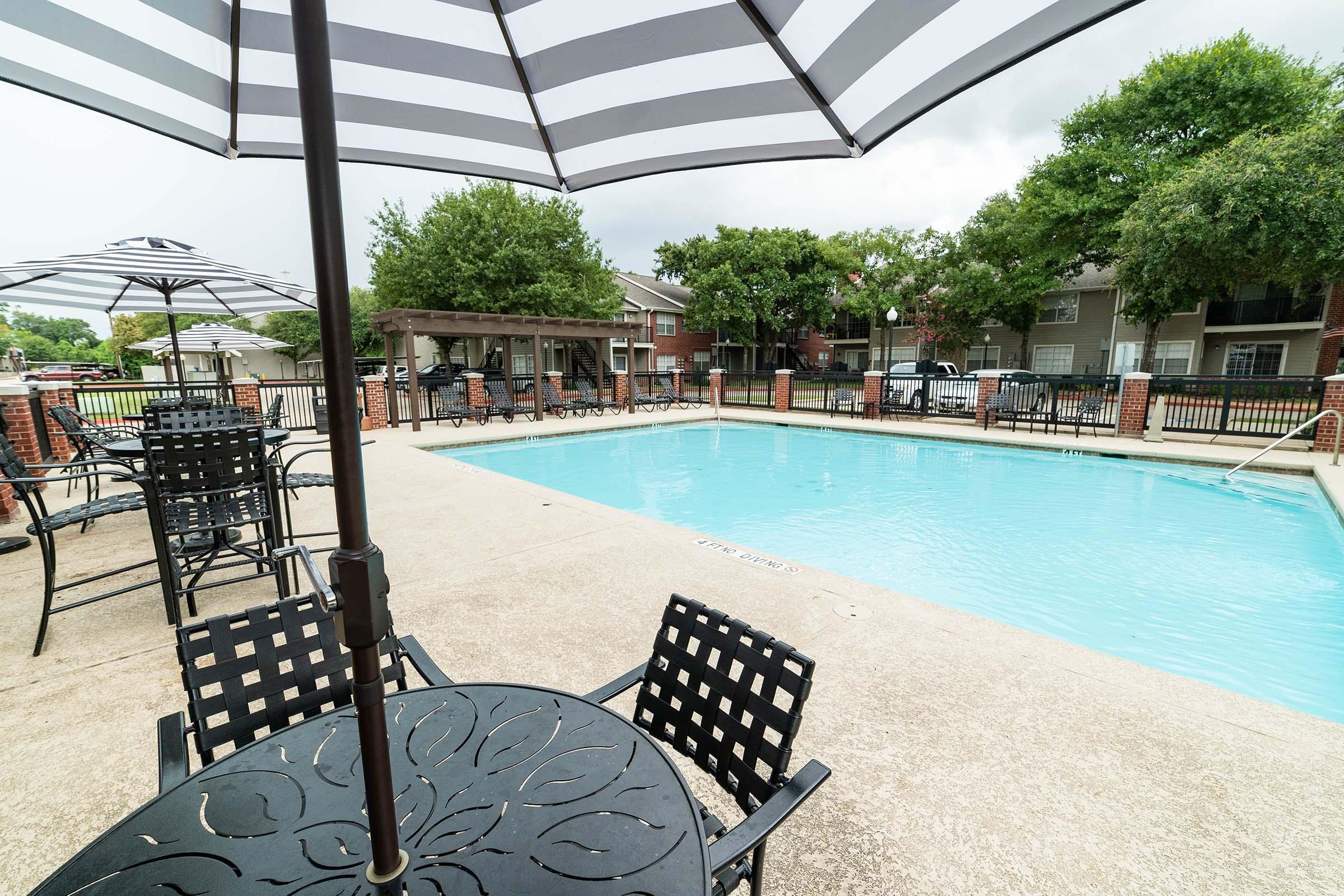 A clear blue swimming pool surrounded by a concrete deck, with several black metal tables and chairs. There are umbrellas providing shade, and a covered seating area in the background. The scene is set in a residential area with green trees and buildings visible nearby, under a cloudy sky.