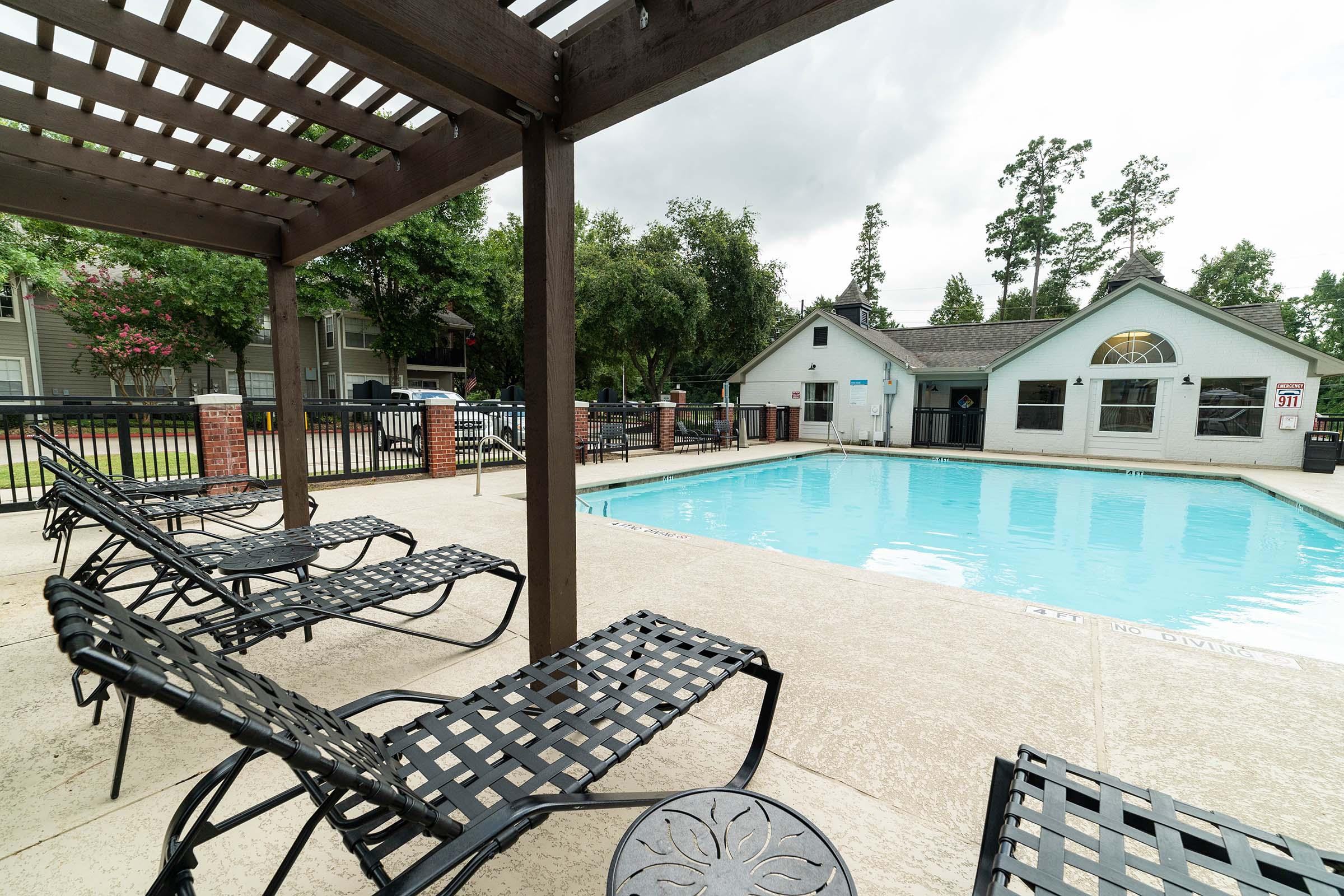 A view of a residential pool area featuring a clear blue swimming pool surrounded by lounge chairs. There is a shaded pergola on one side, and the background includes trees and a building with windows. The scene is inviting and suitable for relaxation.