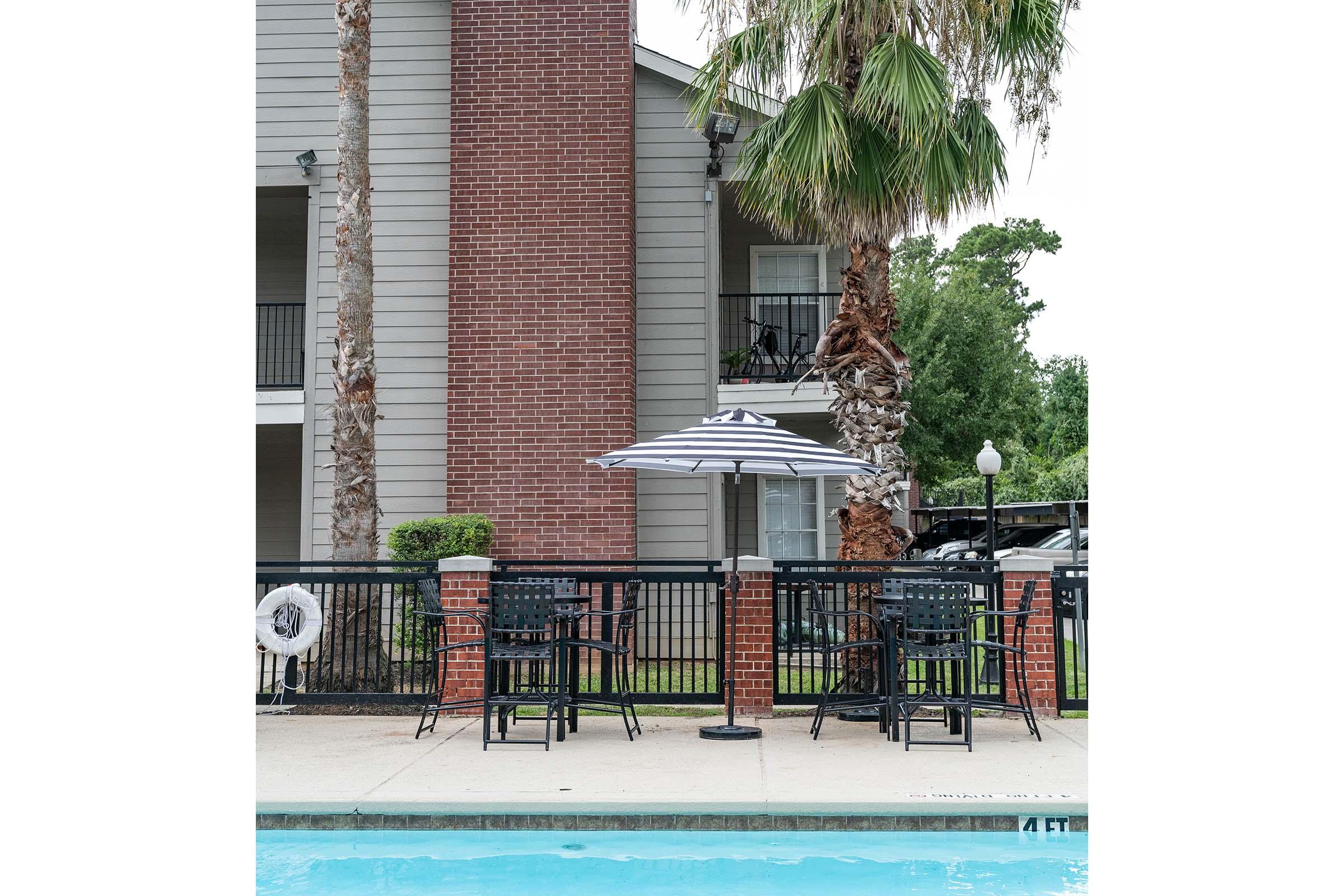 A poolside view featuring black patio furniture, including chairs and a table under a striped umbrella. In the background, there is a multi-story apartment building with palm trees and a railing near the pool area. A lifebuoy is hanging on the fence by the pool.