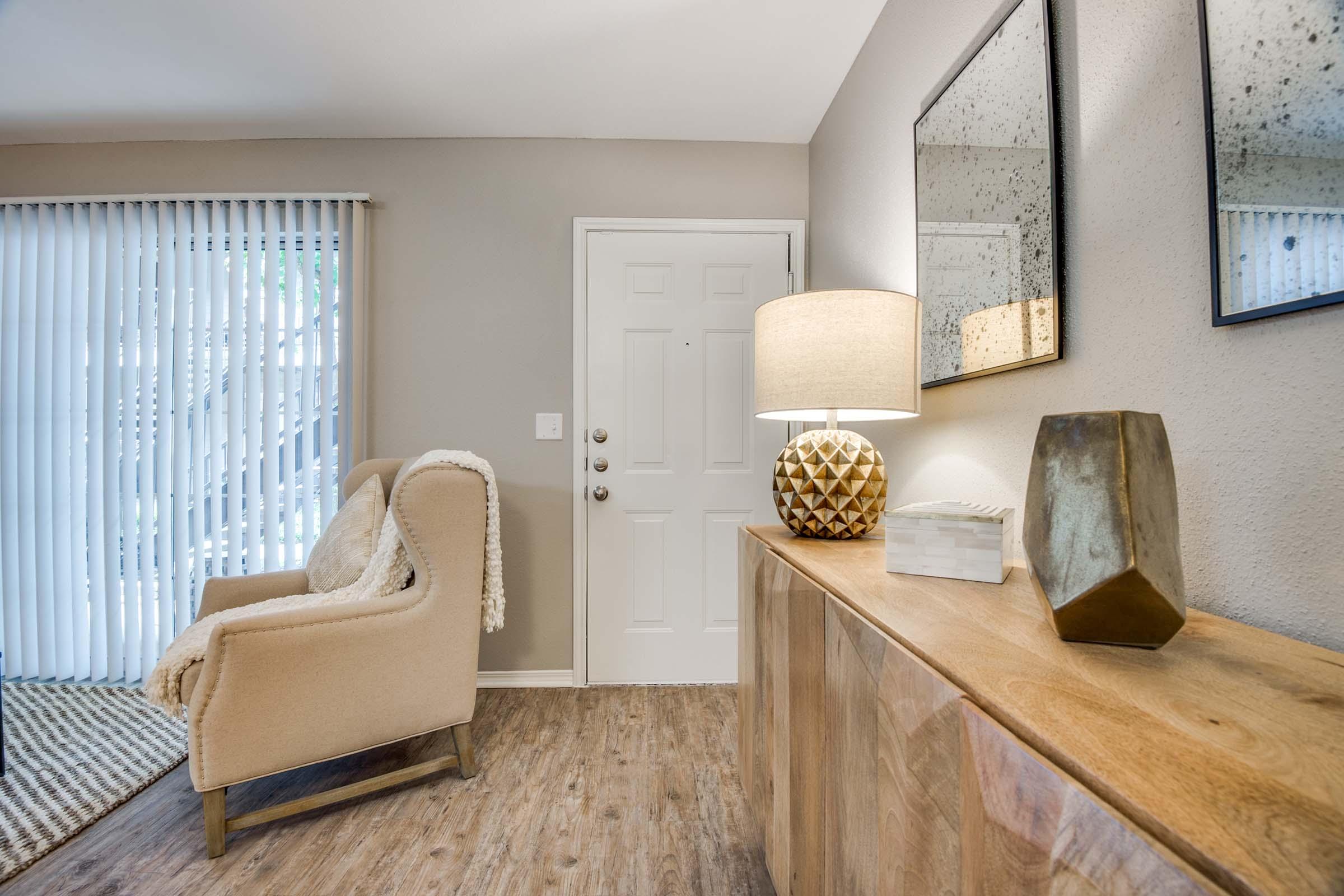 A cozy entryway featuring a light-colored armchair next to a wooden console table. On the table sits a decorative lamp, a geometric vase, and a small box. The wall has two mirrors reflecting the space, and a door with a white blind is visible in the background, enhancing the warm, inviting atmosphere.