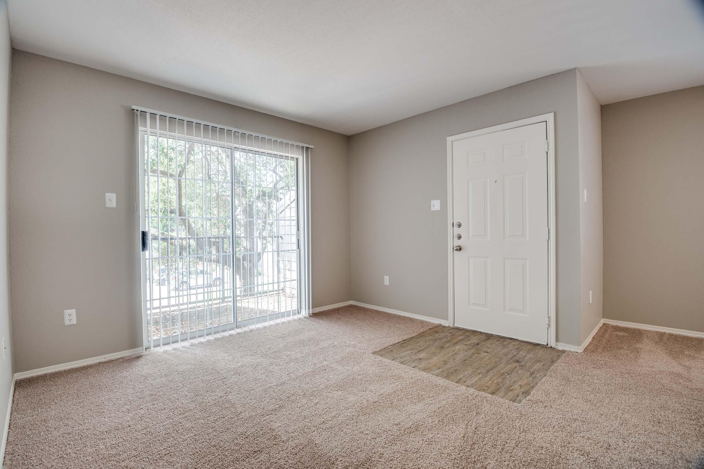 Spacious living room with neutral-colored walls and plush carpet, featuring a sliding glass door that leads to a balcony, allowing natural light to fill the space. The front door is visible on the right side, enhancing the room's inviting atmosphere.