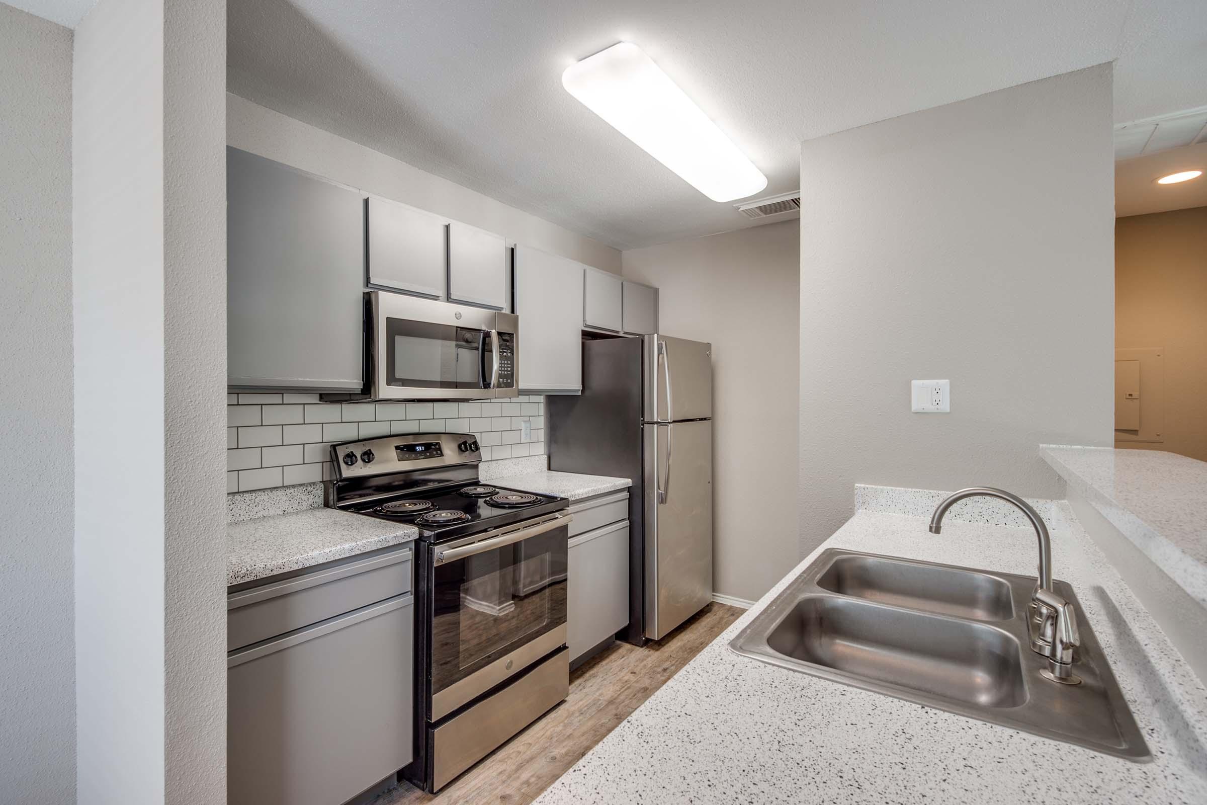 Modern kitchen featuring light gray cabinets, stainless steel appliances including an oven, microwave, and refrigerator. The countertops are a light-colored granite, and the backsplash is white subway tile. A double sink is visible with a sleek faucet, and the space is well-lit by overhead fluorescent lighting.