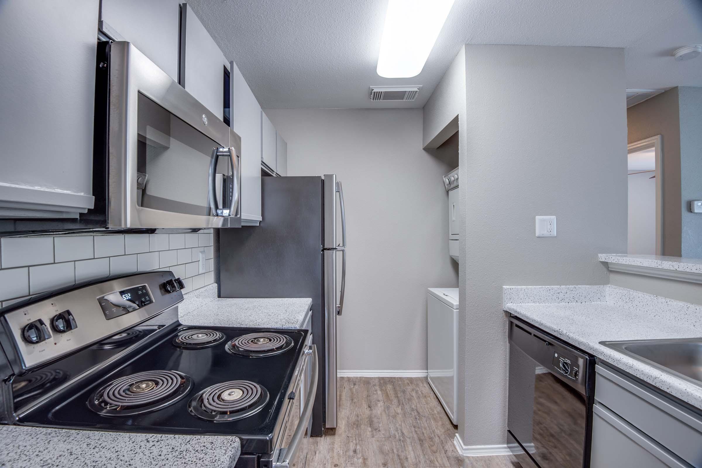 Modern kitchen featuring stainless steel appliances, including a microwave, stove, and fridge. The layout includes gray walls, a granite countertop, and a decorative tile backsplash. There’s also a washer and dryer in the corner, with wood-style flooring adding warmth to the space.