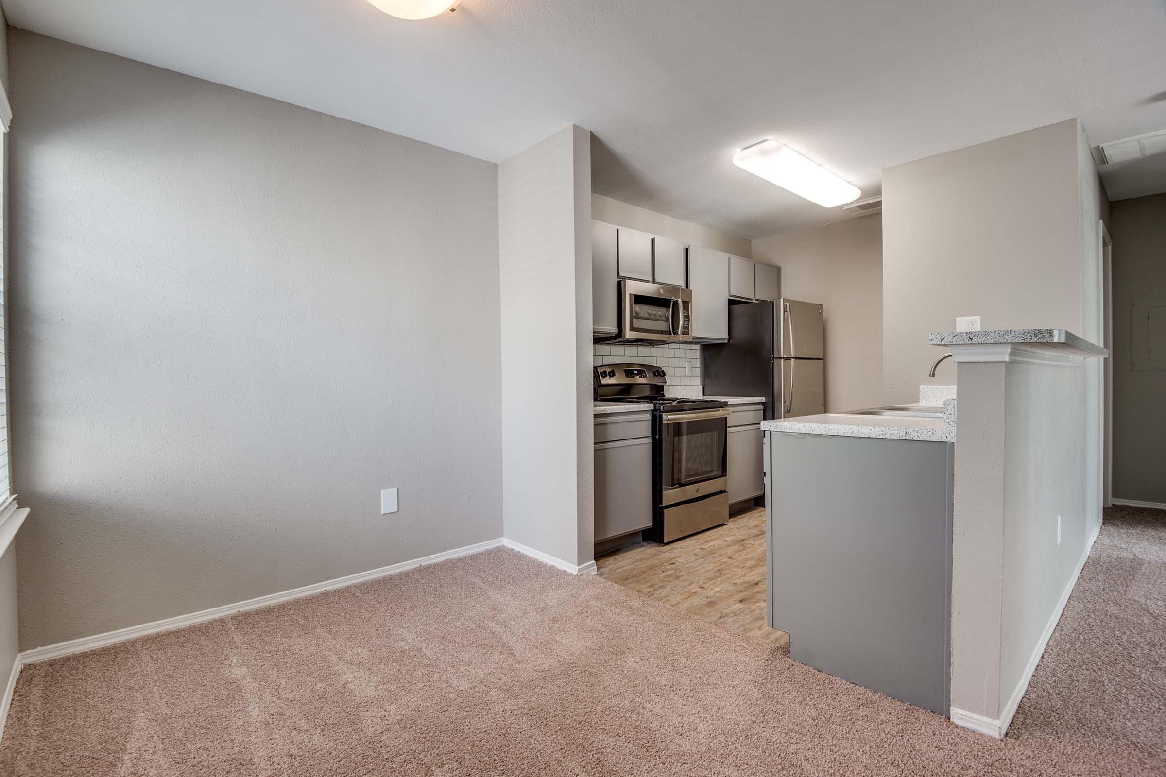 A well-lit, modern kitchen and living area with gray walls and plush carpeting. The kitchen features stainless steel appliances, including a stove and refrigerator, along with light-colored cabinets. A bar-style countertop separates the kitchen from the living space, creating an open-concept feel.