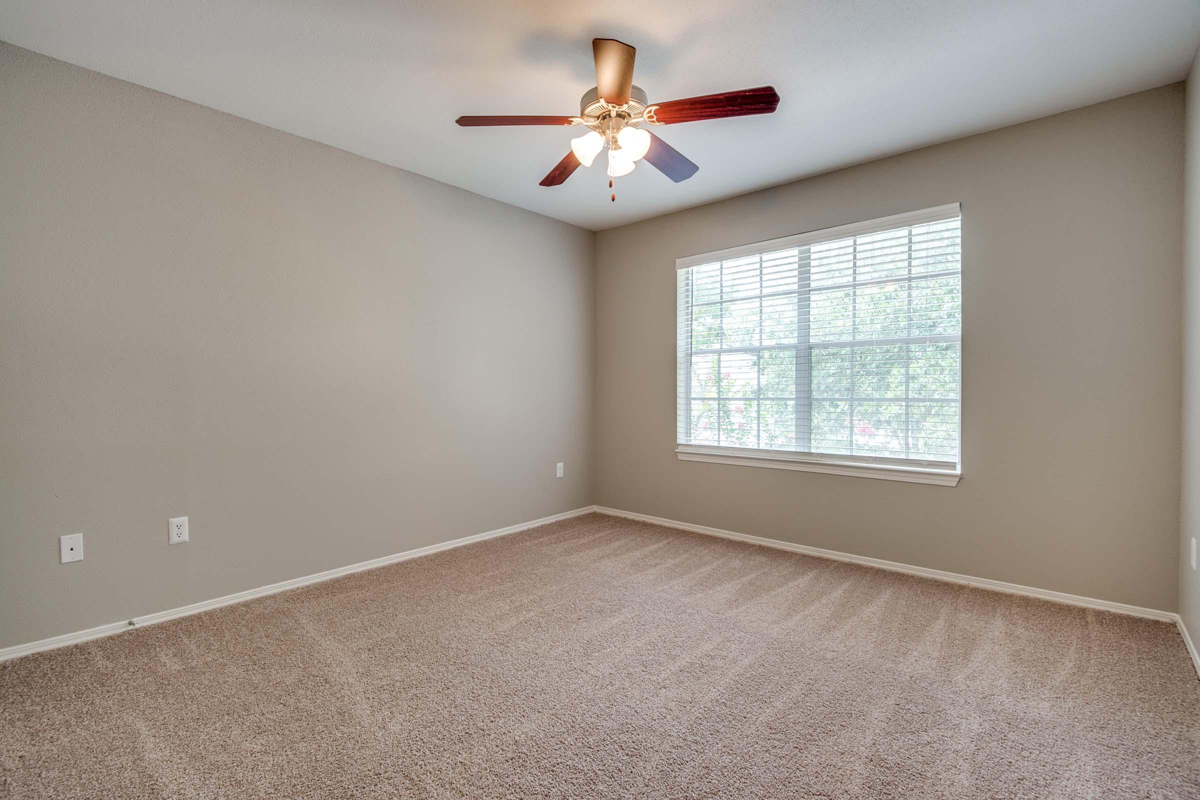 A spacious, empty bedroom featuring beige walls and a large window with white blinds. The room has light brown carpet and a ceiling fan with wooden blades. Natural light floods in, illuminating the space, which is ready for furniture and personal touches.