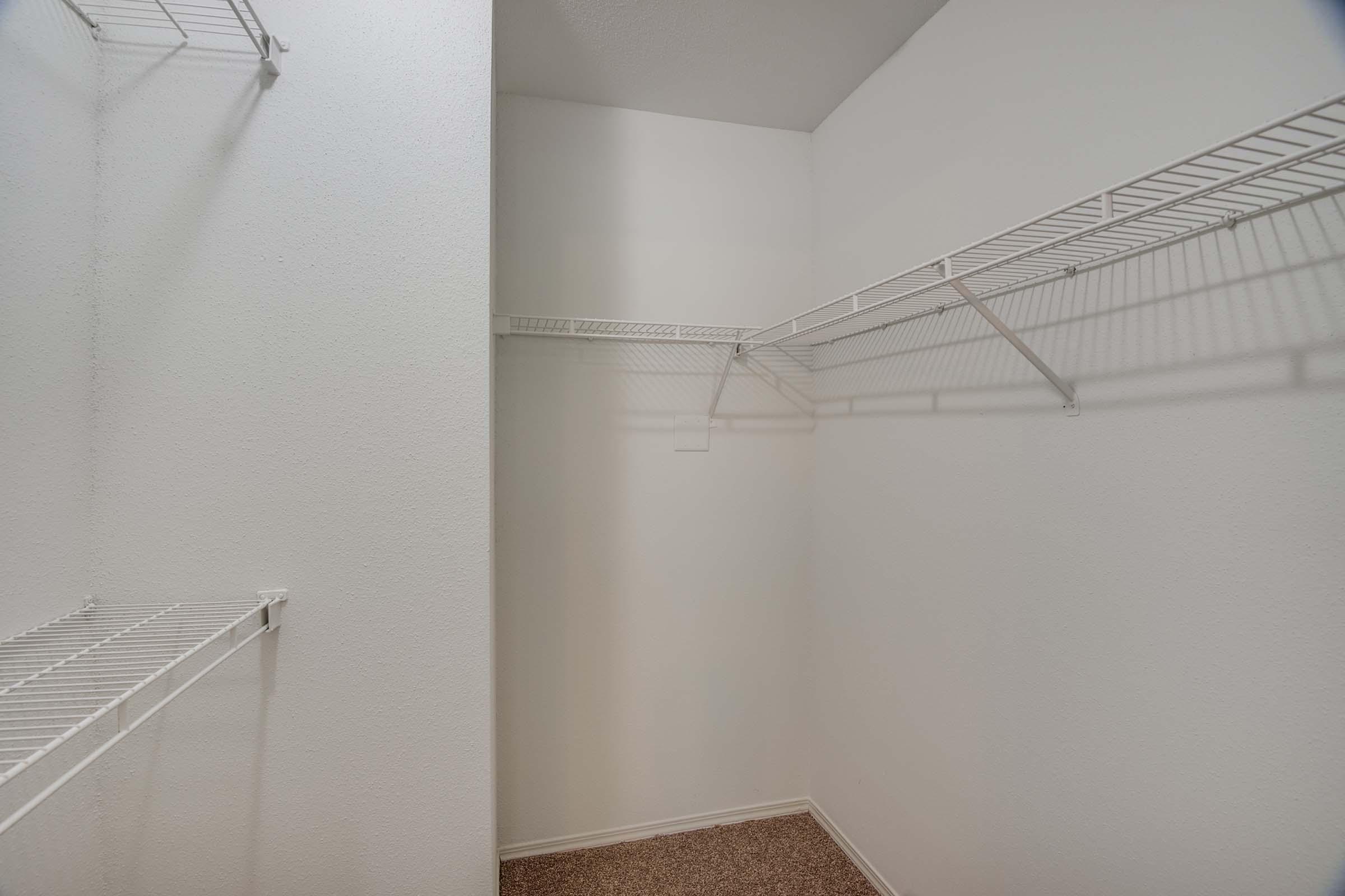 Empty closet with white walls and metal wire shelving. One side has a single shelf, and the other side shows a larger section with two shelving units, all against a light-colored wall. The floor is covered with tan carpeting. Natural light illuminates the space.
