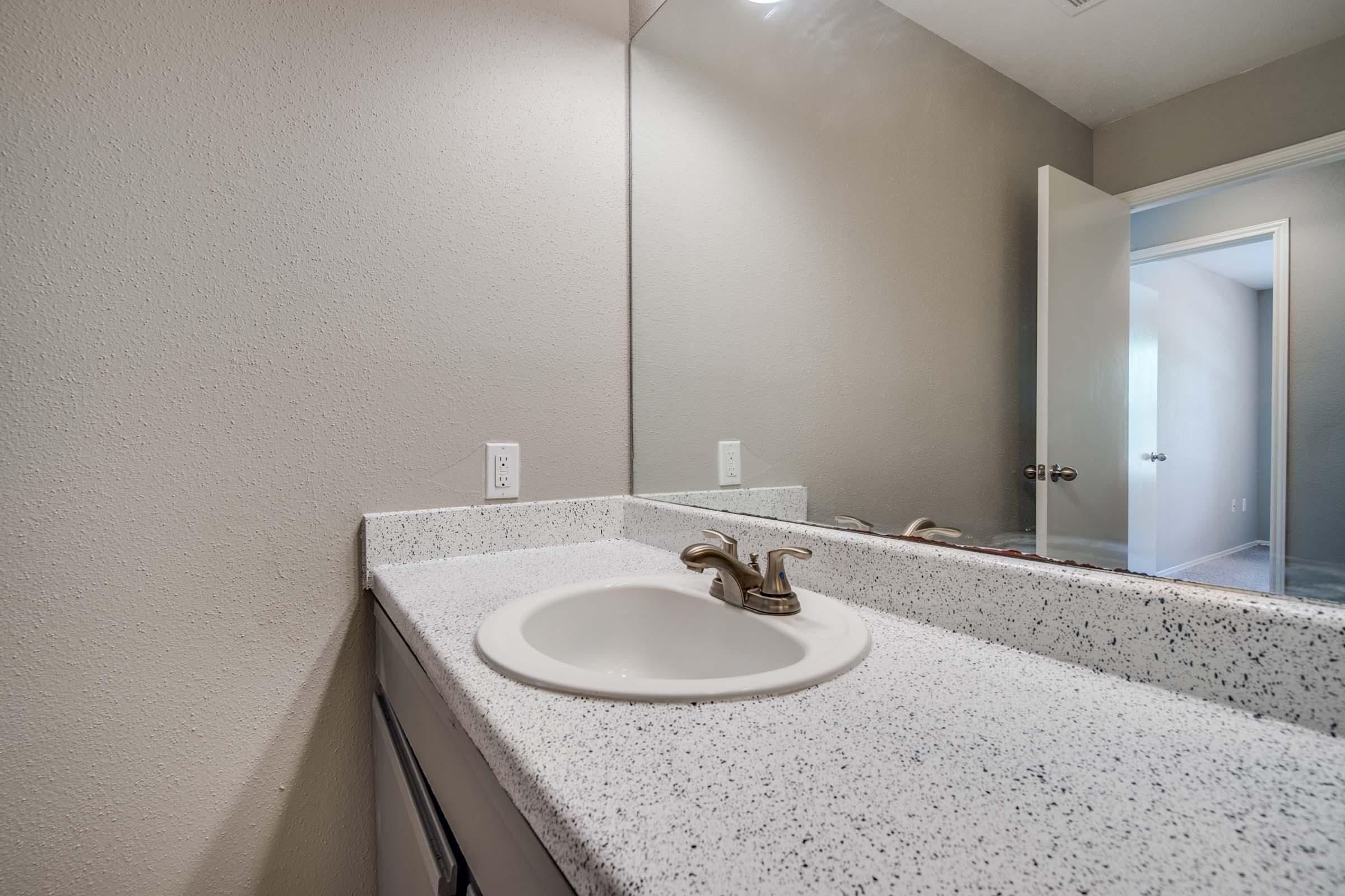 A clean bathroom sink area with a speckled countertop, featuring a faucet, a wall mirror, and an open door leading to another room. The walls are painted a light color, providing a bright and airy atmosphere.