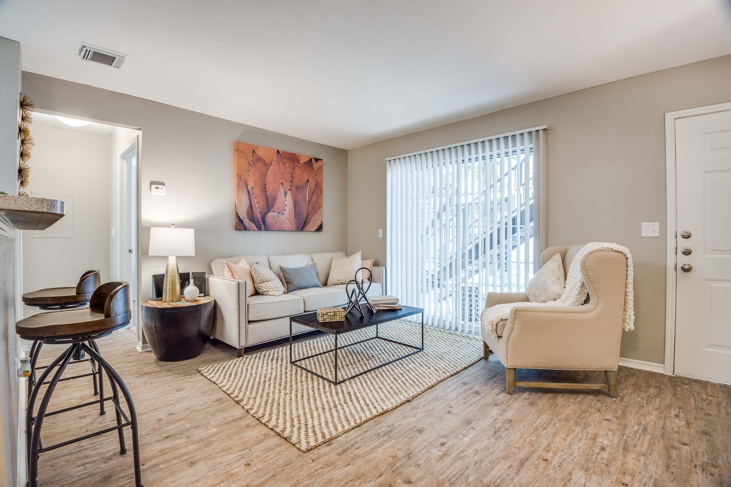 A cozy living room featuring a light gray sofa with decorative pillows, a round coffee table with a decorative centerpiece, and a beige armchair. A textured area rug adds warmth to the wooden floor. There are sliding glass doors with vertical blinds allowing natural light, and a modern art piece on the wall.