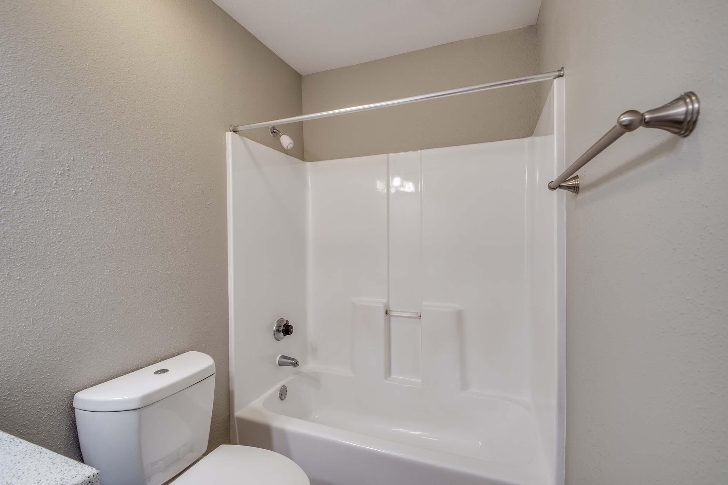 A clean bathroom featuring a white tub and shower combination with a silver curtain rod. There is a modern toilet and a light-colored countertop visible. The walls are painted a neutral tone, providing a simple and fresh aesthetic.