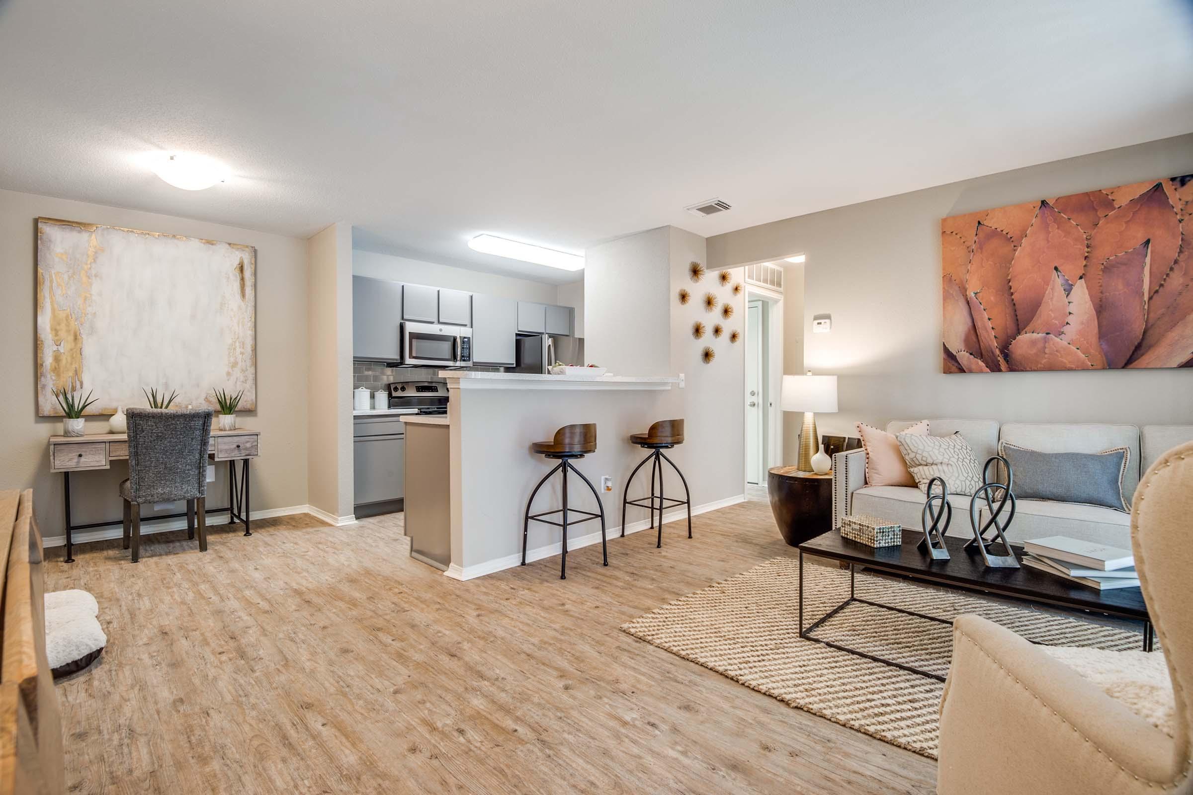 A modern and stylish living room featuring a beige sectional sofa, a black coffee table, and a woven area rug. The open layout includes a dining area with bar stools, a kitchenette visible in the background, and decorative wall art, creating a warm and inviting atmosphere.