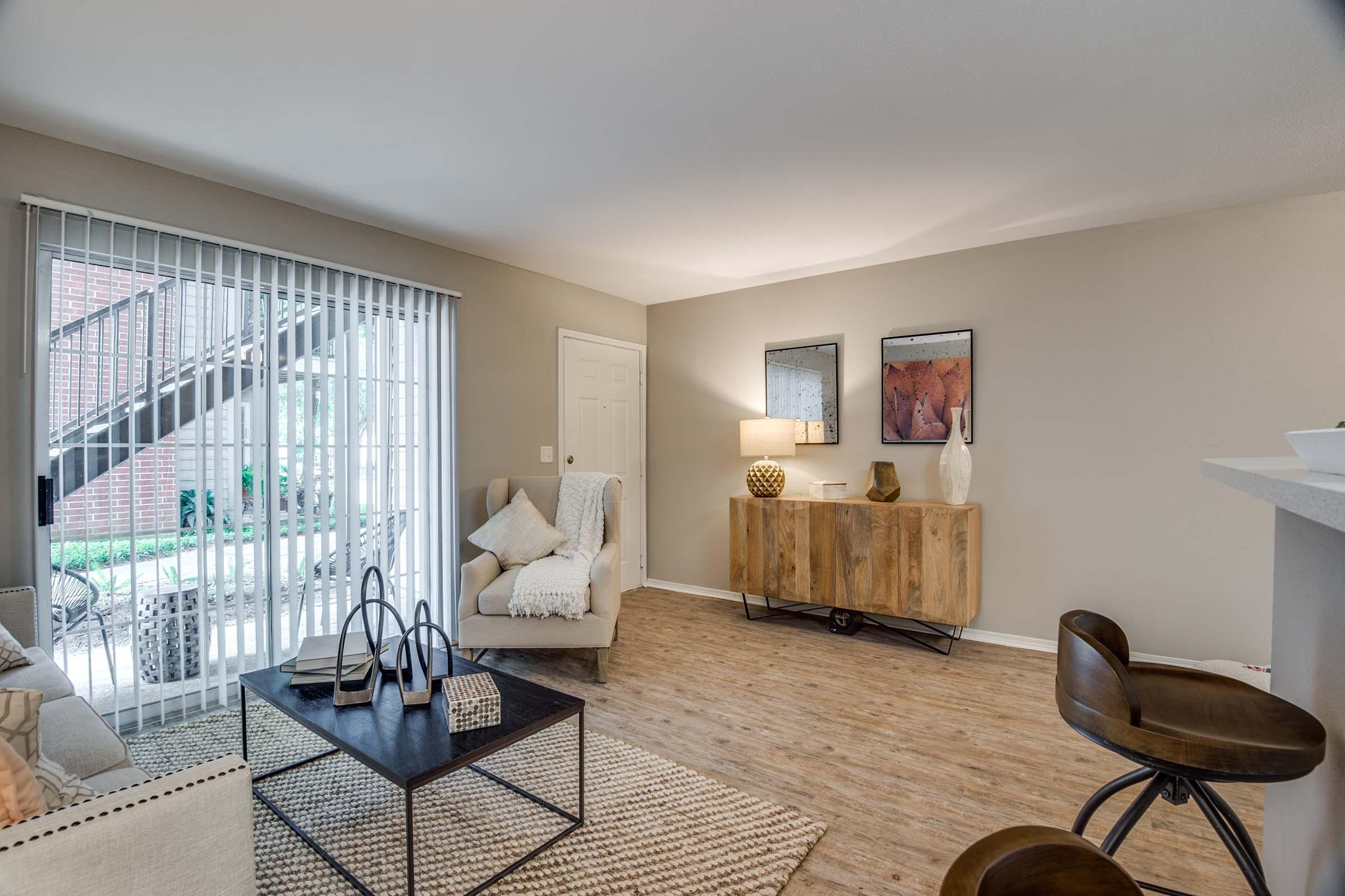 A cozy living room with light-colored walls and wooden floors. It features a cream armchair with a decorative throw, a black coffee table, and a stylish wooden sideboard. There are artworks on the walls, and large sliding doors lead to an outdoor space. Natural light fills the room, creating a warm atmosphere.