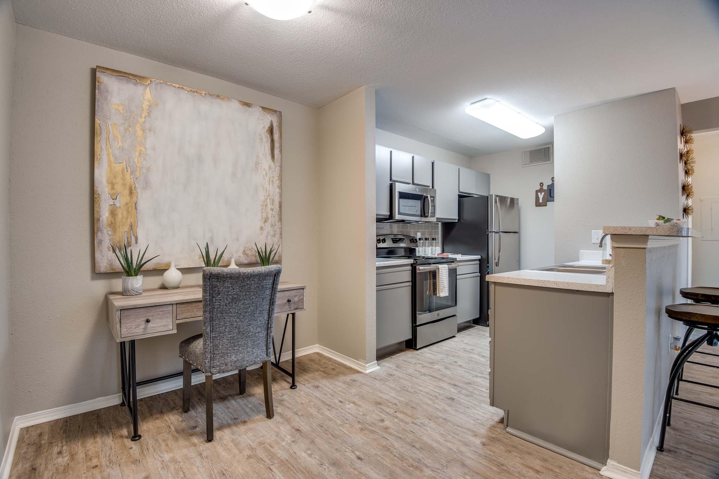 Modern kitchen and dining area with a sleek design. The kitchen features stainless steel appliances and light-colored cabinetry. A small desk with a gray chair is positioned near the wall, adorned with a large abstract art piece and decorative plants. The flooring is light wood, creating a warm atmosphere.