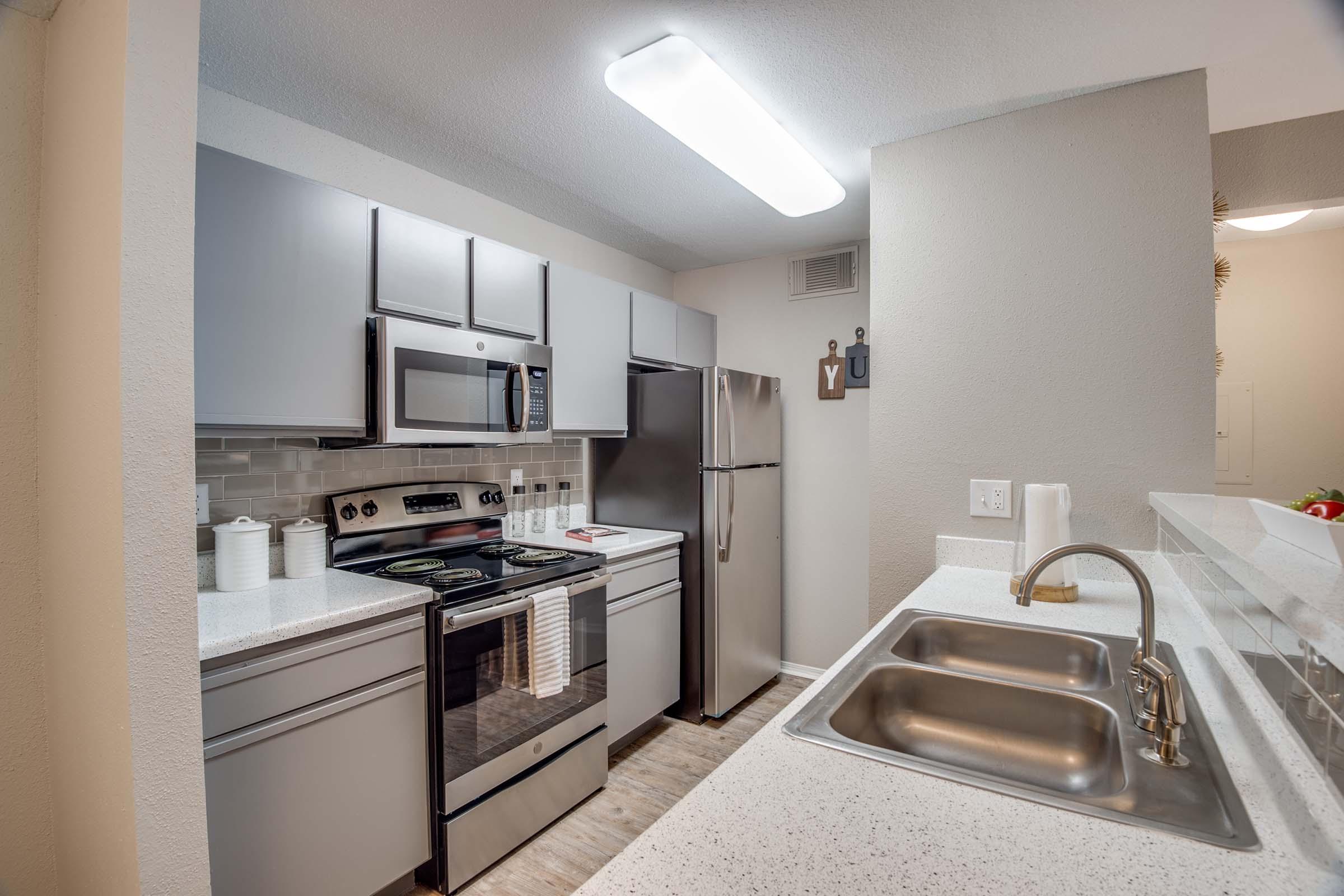Modern kitchen with gray cabinetry, stainless steel appliances including a microwave and refrigerator, and a double sink. The countertop is white with a speckled design. Brightly lit space with a light fixture above, tiled backsplash, and a few kitchen accessories on display.