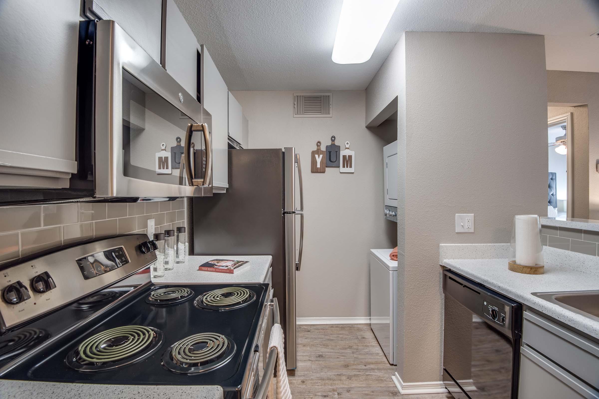 A modern kitchen featuring stainless steel appliances, including a stove, refrigerator, and microwave. The countertop is white with a subtle texture, and the light-colored cabinets complement the neutral wall color. A small laundry area is visible in the adjacent space. Decorative letters spell "YUM" on the wall.