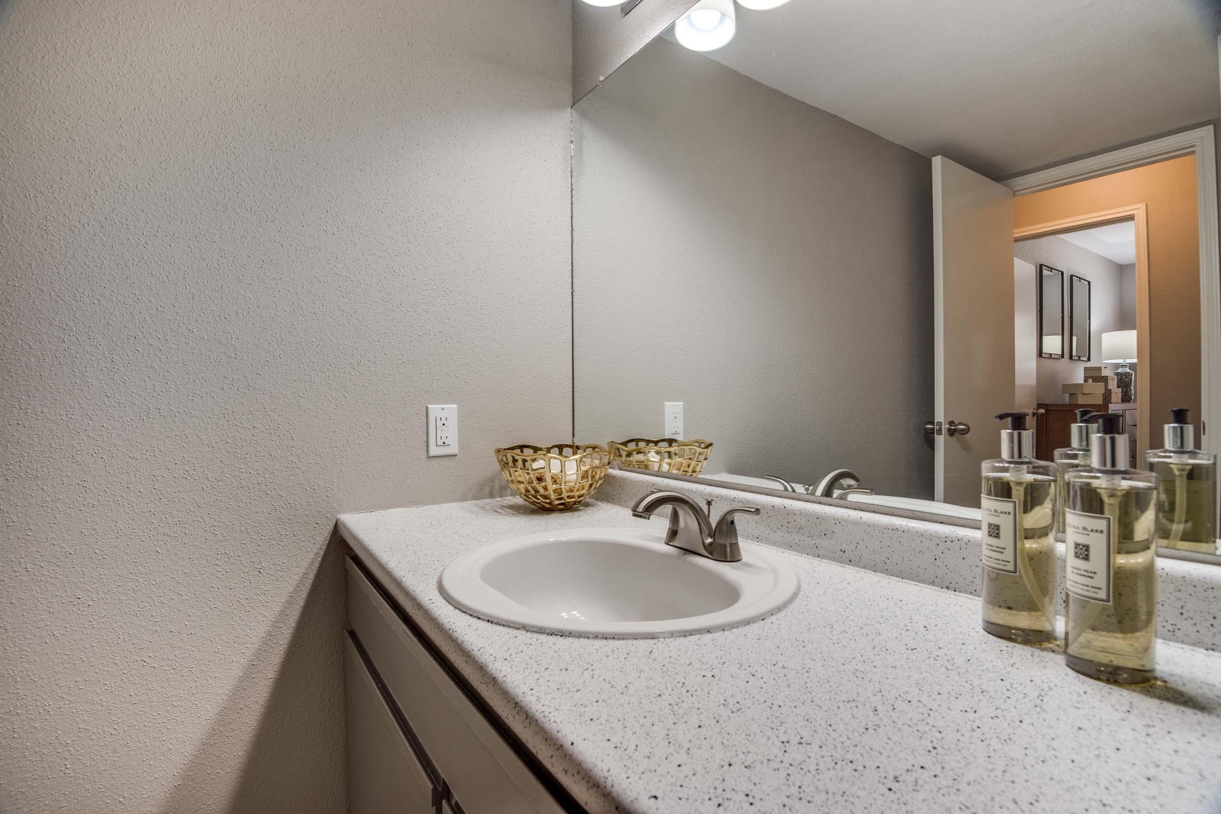 A modern bathroom countertop with a white sink, decorative gold bowl, and glass bottles of soap. The walls are painted in a neutral shade, and there's a mirror illuminated by overhead lights. A doorway leads to another room in the background.