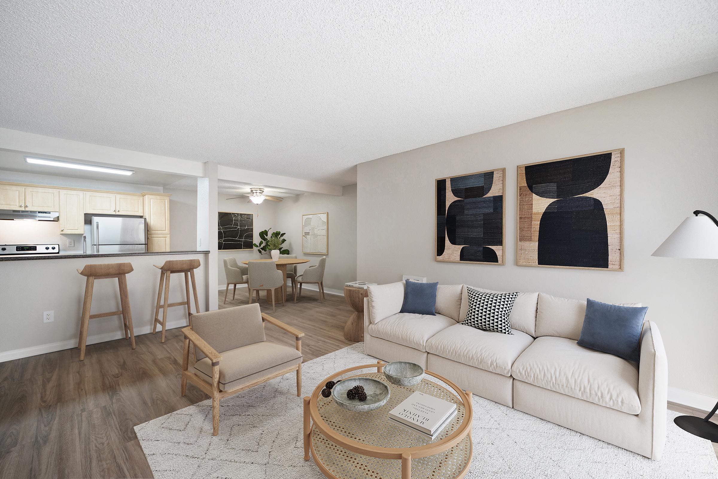 A modern living room with light-colored walls and a comfortable sofa featuring decorative pillows. There’s a round coffee table with a glass top, wooden accent chairs, and abstract wall art. In the background, a kitchen area is visible, furnished with wooden bar stools and dining space. Warm, inviting decor enhances the ambiance.