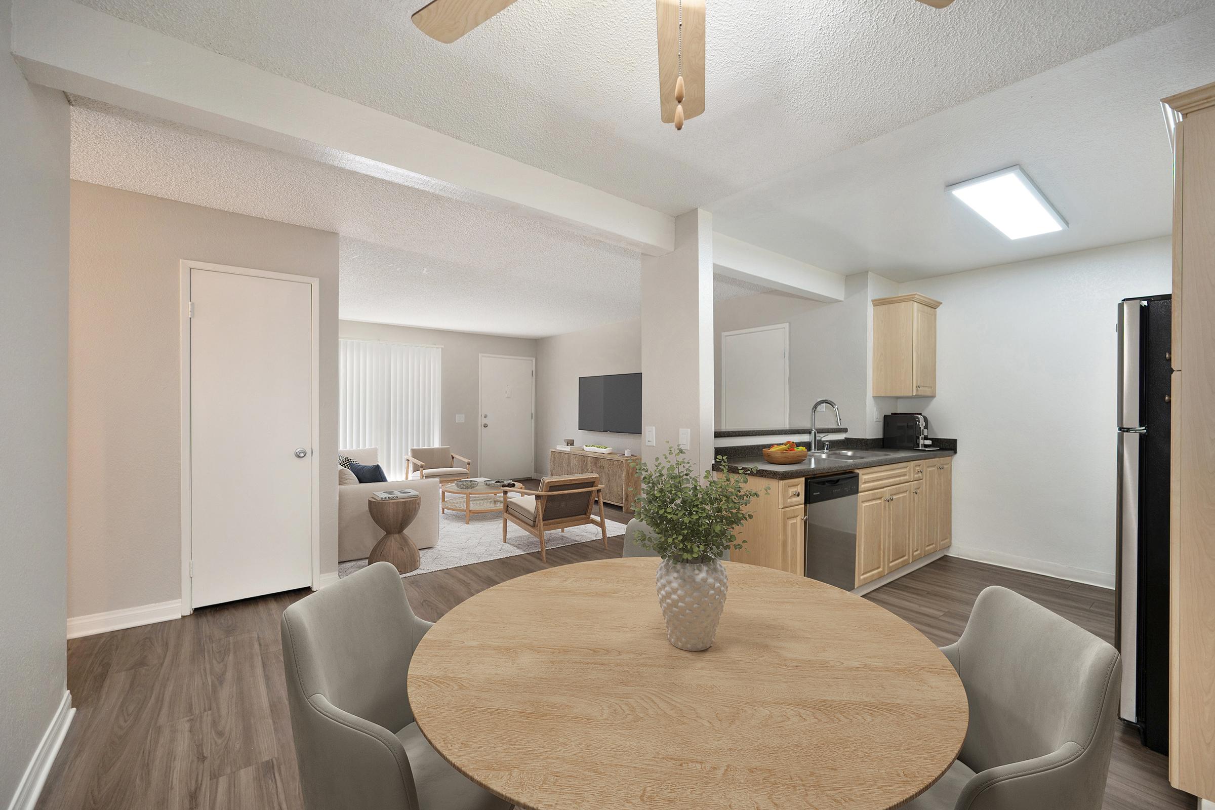 A modern kitchen and dining area featuring a round wooden table with light-colored chairs, a kitchen equipped with wooden cabinets, a black refrigerator, and a spacious living room visible in the background, furnished with a sofa and a television. Bright lighting enhances the open and inviting atmosphere.