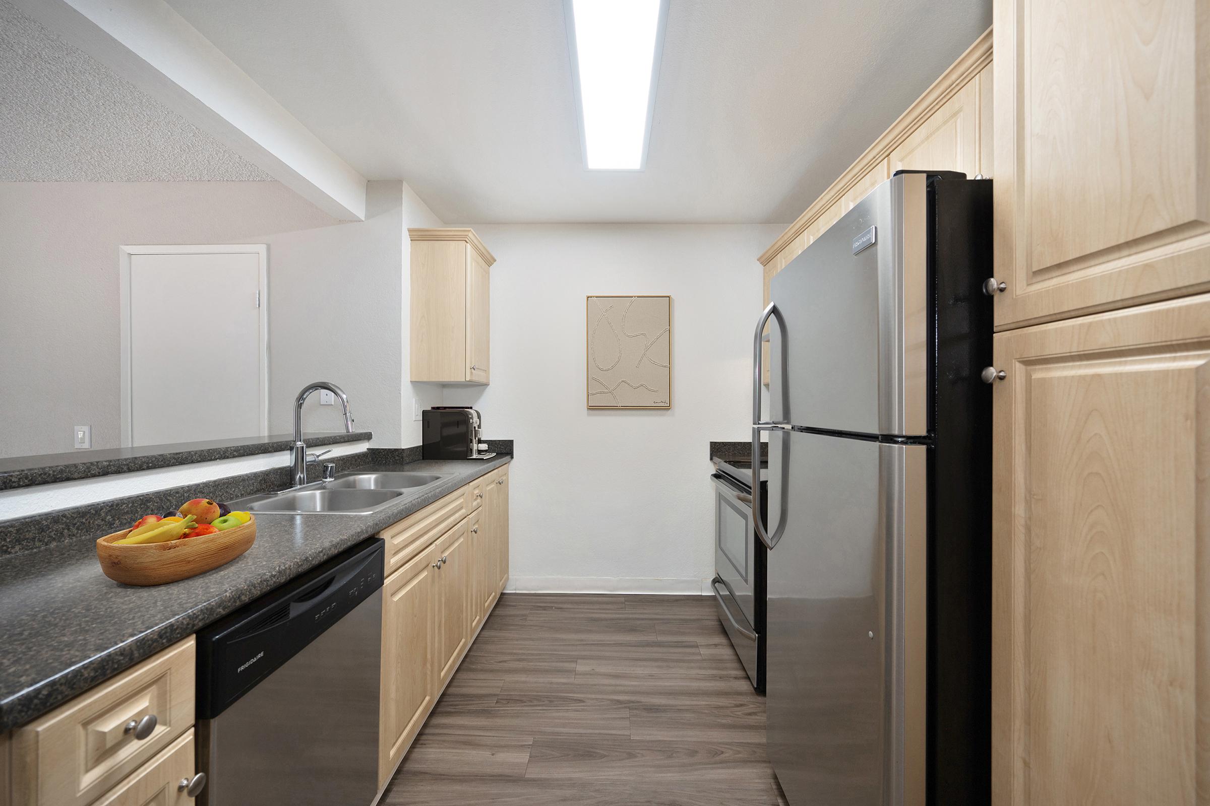 Modern kitchen featuring light wood cabinetry, a stainless steel refrigerator, and a large sink. Quartz countertops are complemented by a smooth backsplash. A fruit bowl filled with colorful fruits is on the left, and a minimalist artwork is mounted on the wall. The space is well-lit with a ceiling light.
